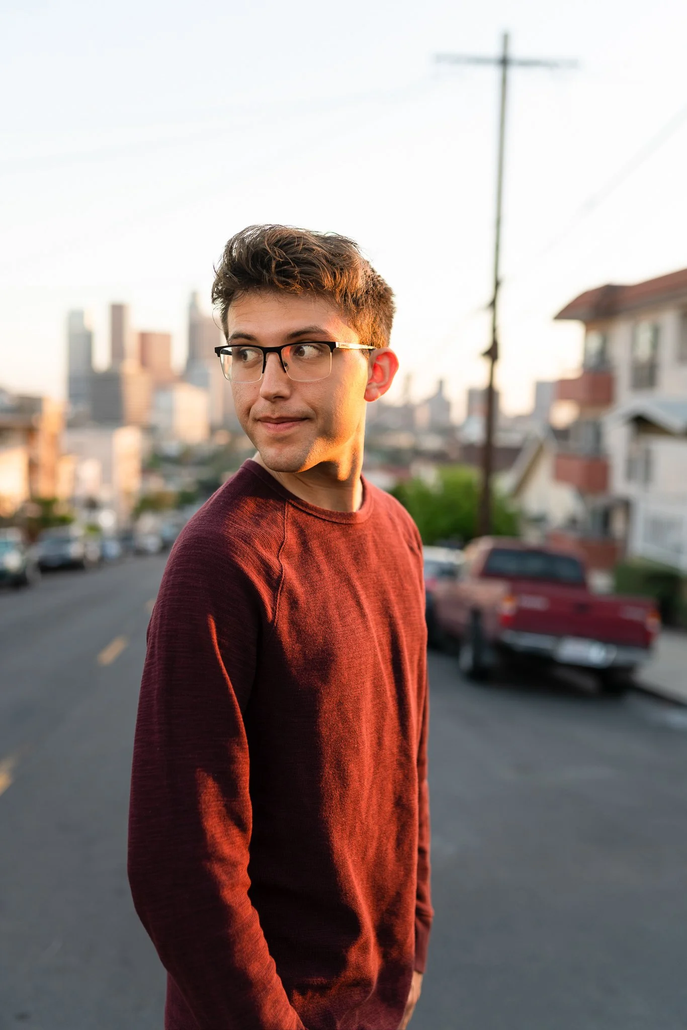 A young man with glasses and brown hair is standing on a street during sunset, looking to his left, with city buildings in the background.