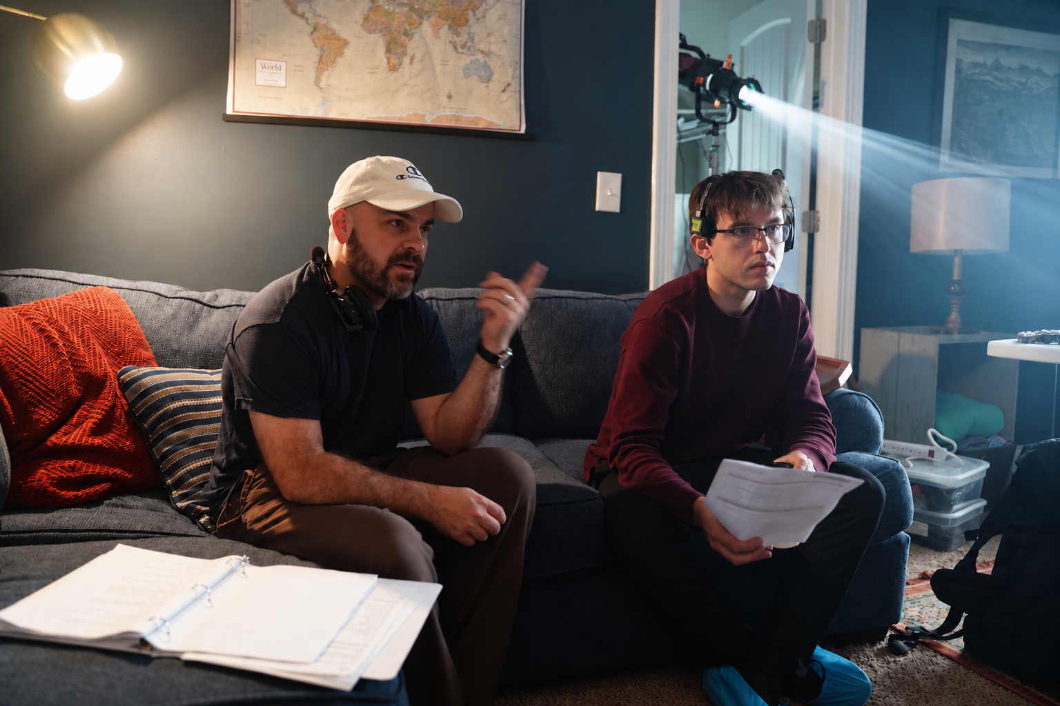 Two men sitting on a dark blue couch in a living room, with one man using a headset and a laptop and the other pointing and speaking, with film equipment and papers around.