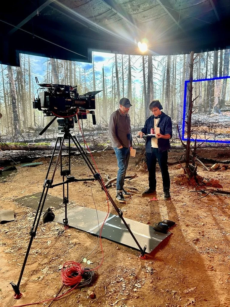 Behind the scenes of a film or TV production set in a forest, featuring a camera on a tripod, two crew members discussing, a light on the ceiling, tools on the ground, and a marked area around a bare tree.