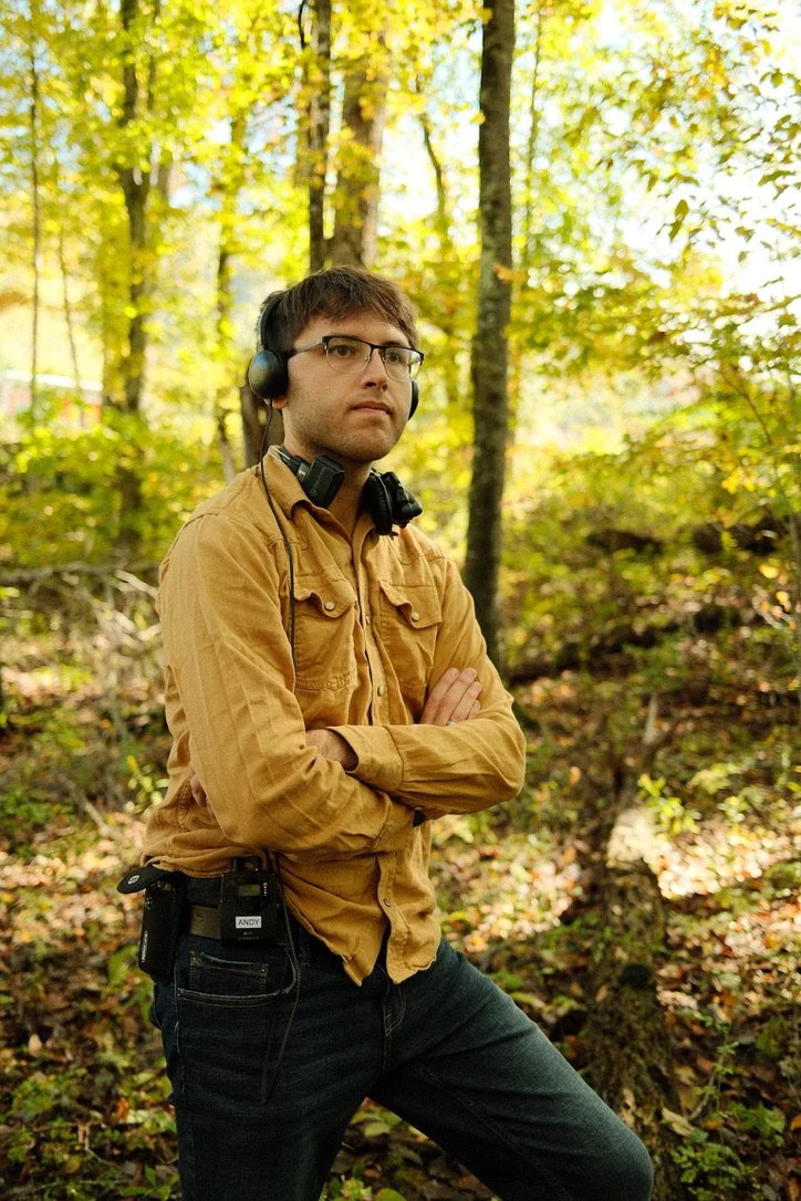 A man with glasses wearing a brown shirt and headphones stands in a forest with arms crossed.