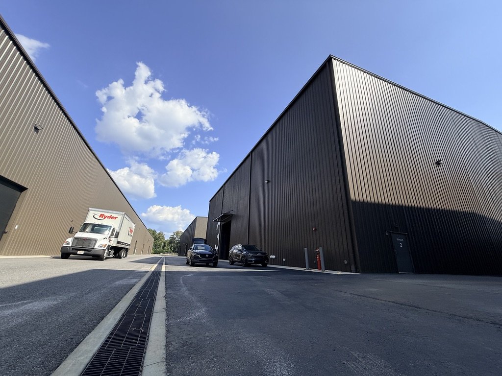 Industrial warehouse buildings with parked cars and a delivery truck under a partly cloudy sky.