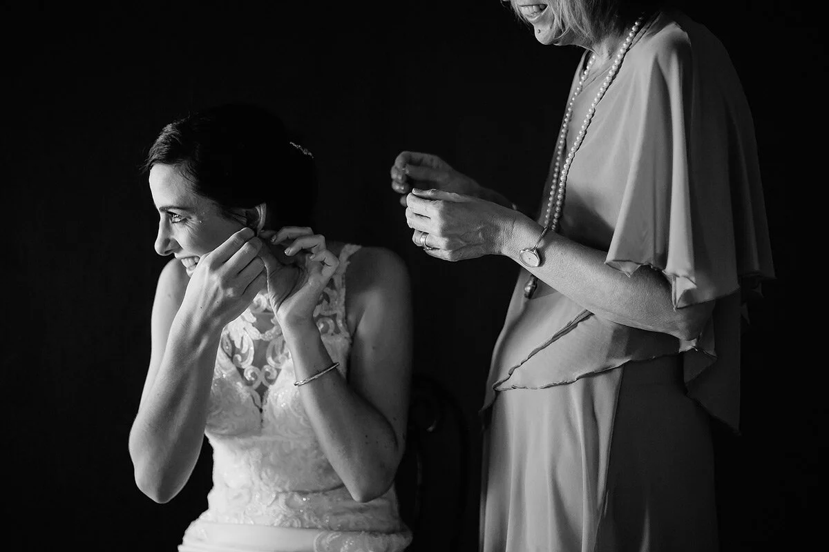 Bride putting on her earrings with her mother in the backround.