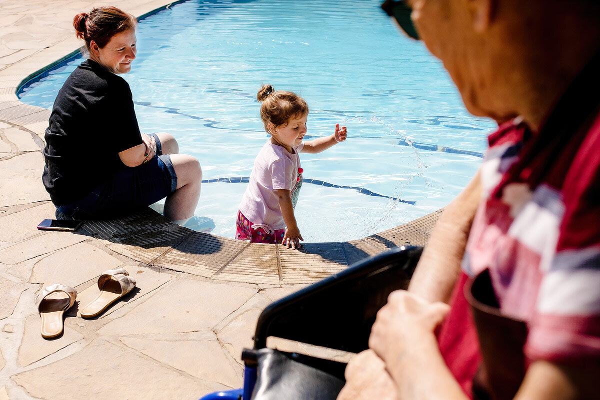 A Kid playing in the swimming pool during a hot wedding day with grandfather and mother looking on.