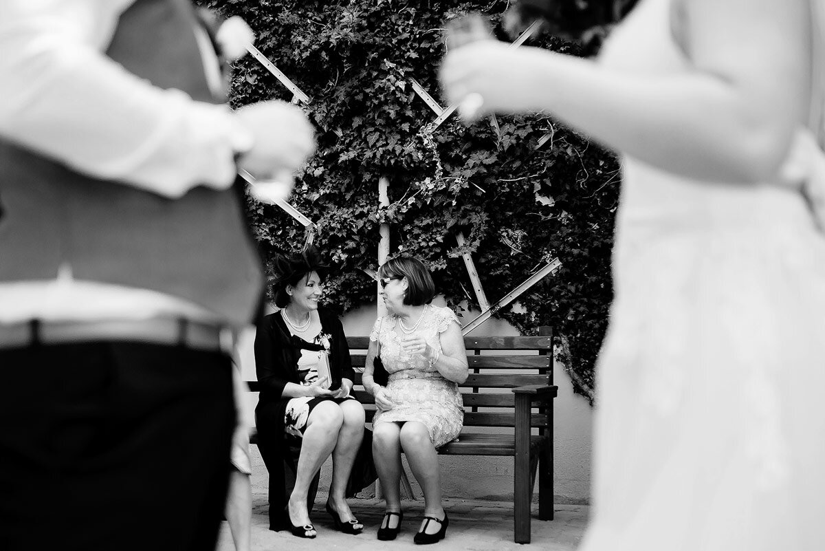 Guests at a wedding enjoying pre-drinks on a bench with bride and groom in the foreground.