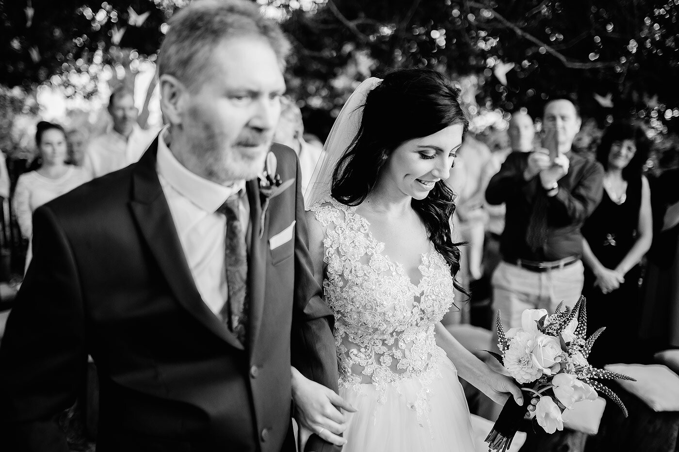 Bride and her father walking down the aisle between oak trees.