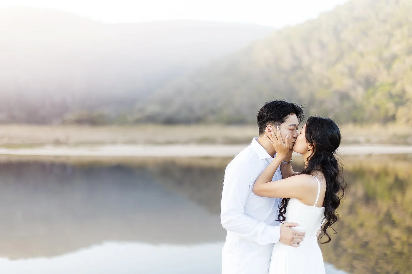 A couple kiss with lake and mountains as backdrop.
