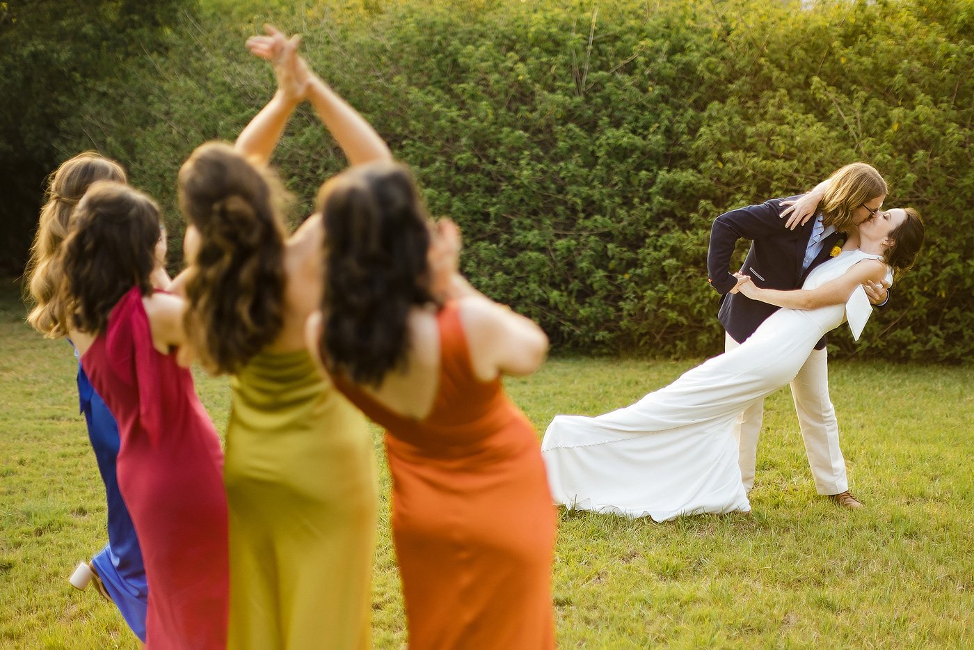 Couple with dip kiss while bridesmaids in colourful dresses looks on.