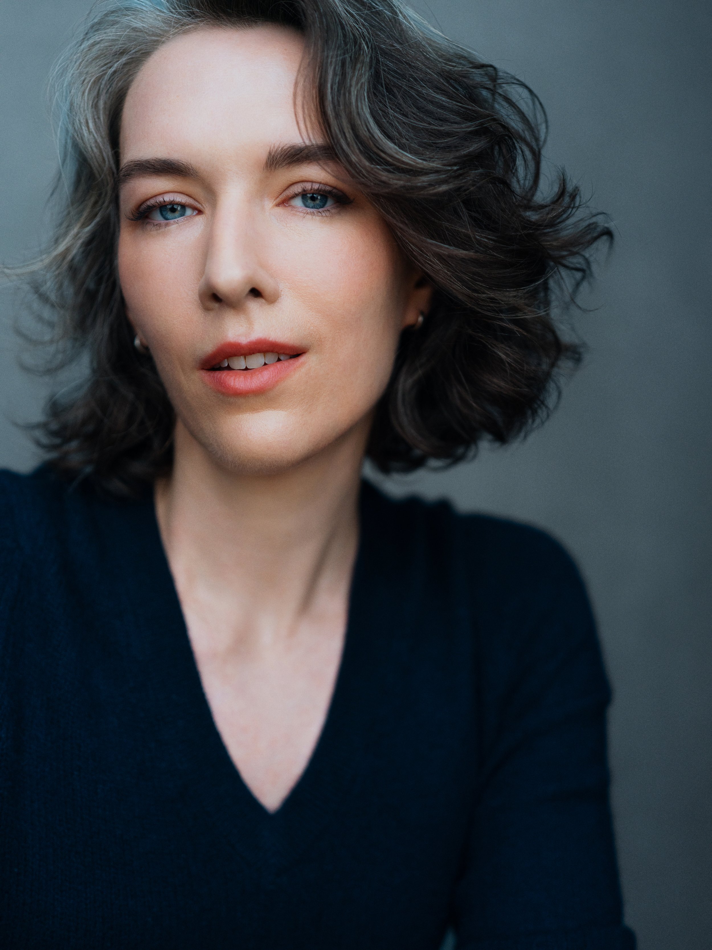 Krystal Riggs, young woman who reads 30s, sits looking into the camera with a soft yet direct expression, parted lips, and brown wavy hair flying loosely around her face. She is in a navy v neck sweater. Portrait by Jeff Mosier.
