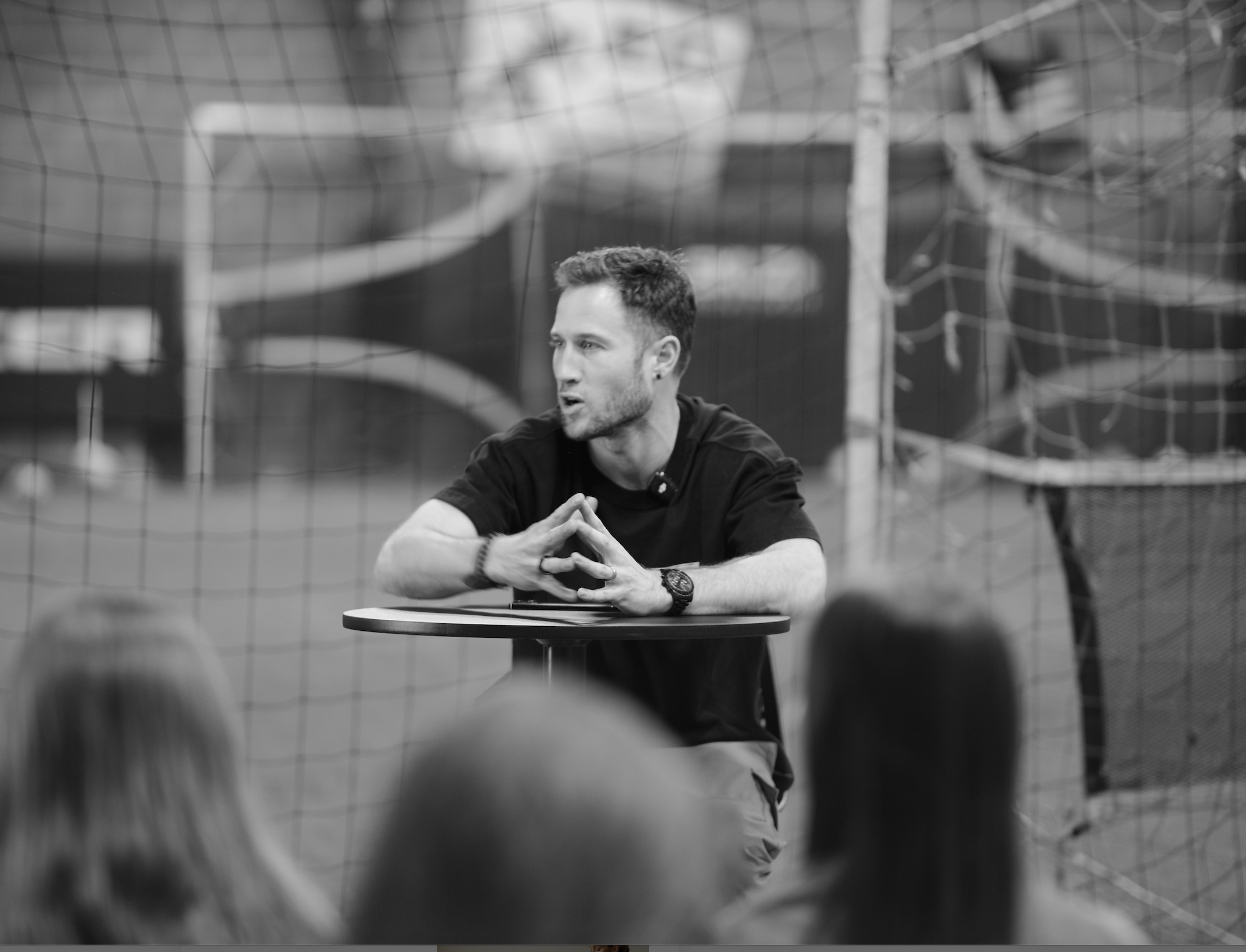 A man with short hair, wearing a black T-shirt, sits at a small table with hands clasped, speaking to an audience in an indoor sports facility with a net in the background.