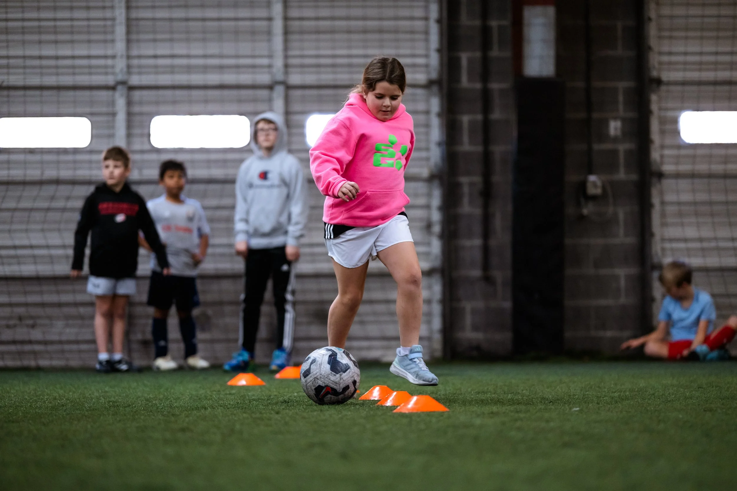 A child in a pink hoodie is playing soccer indoors, dribbling a soccer ball around orange cones. Other children are standing and sitting nearby on artificial turf.