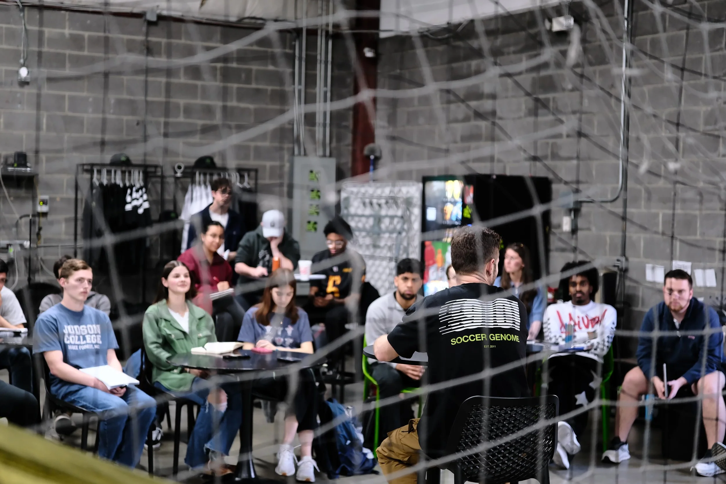 A group of young adults seated in a semi-circle in an industrial-style room with gray brick walls, listening to a person in a black soccer jersey with 'SOCCER GENOME' written on the back, standing at a table.