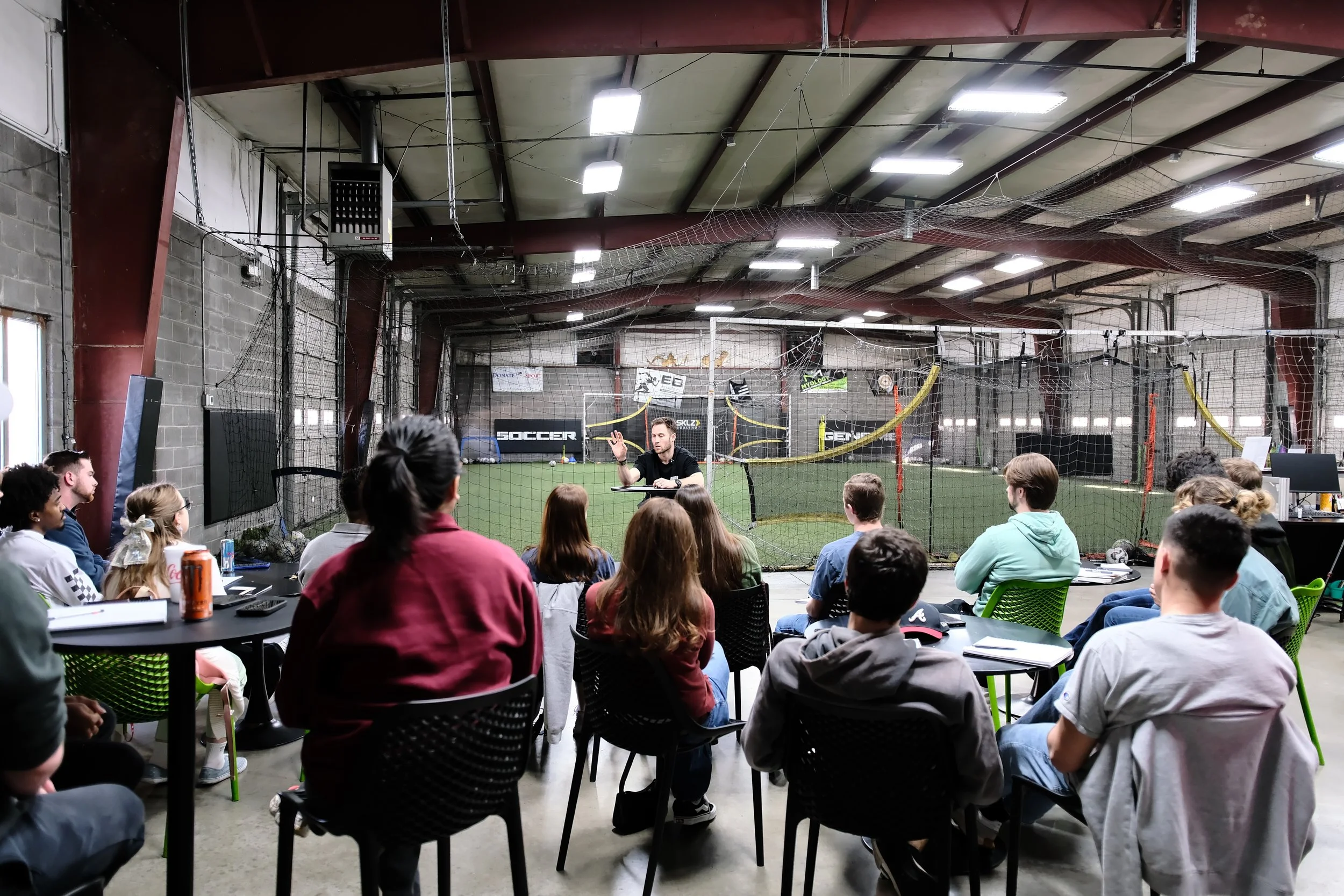 Group of people attending a lecture or seminar inside an indoor soccer facility, with a man presenting at the front and soccer nets and equipment in the background.