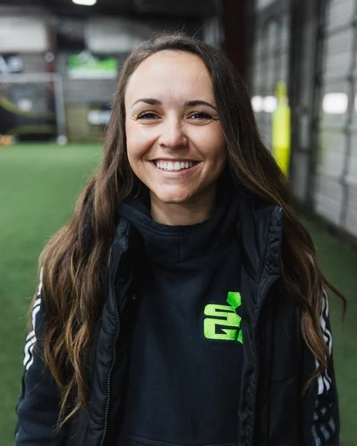 Smiling person in a sports facility, wearing a black jacket and hoodie with a green logo.