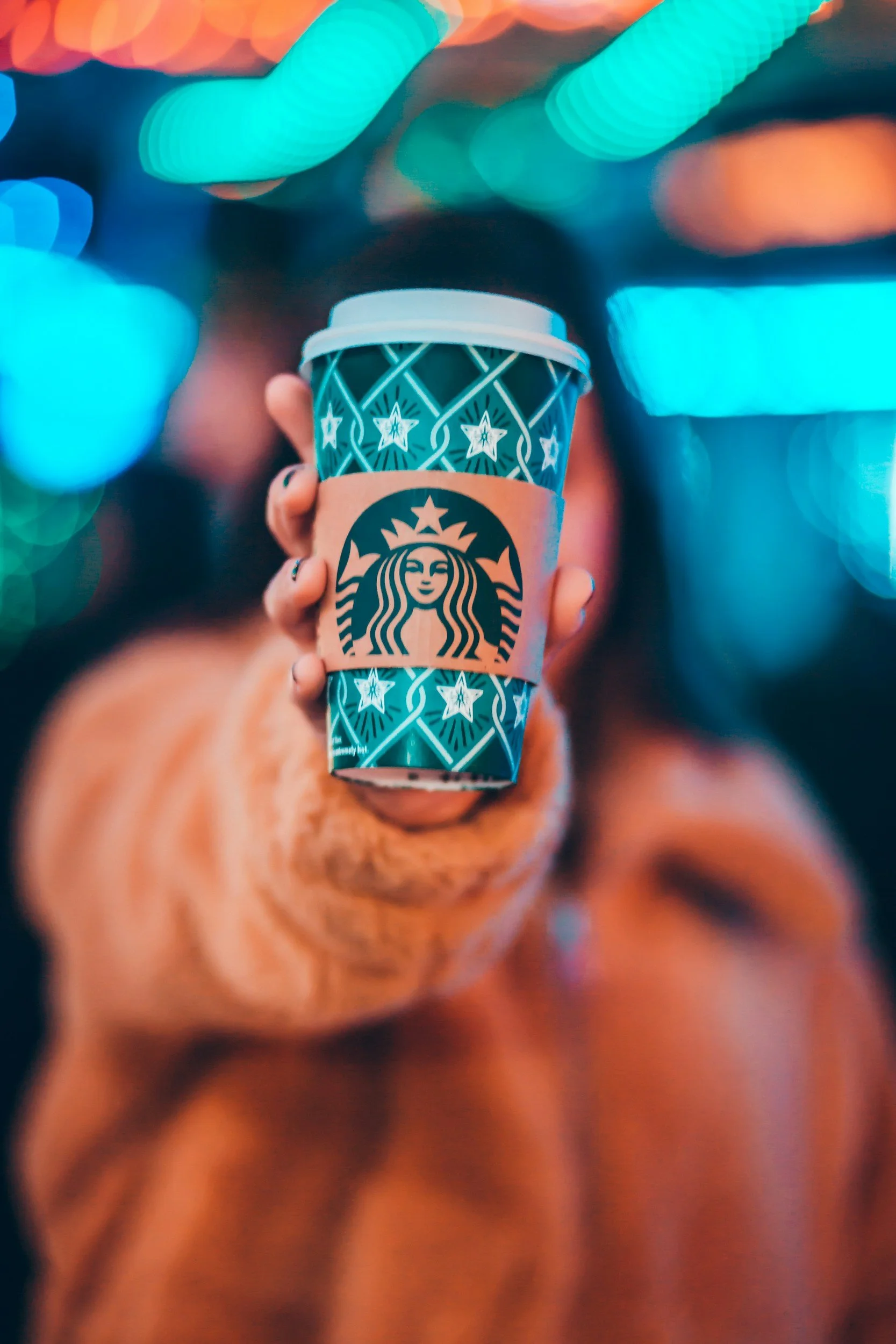 A hand holding a Starbucks coffee cup with a holiday-themed sleeve against a blurred cityscape with colorful lights at night.
