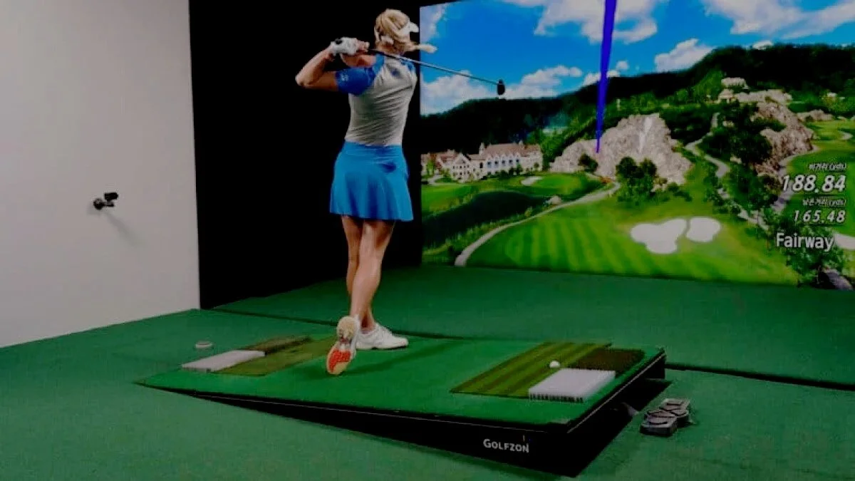 An indoor golf simulator with a woman swinging a golf club on a simulated golf course