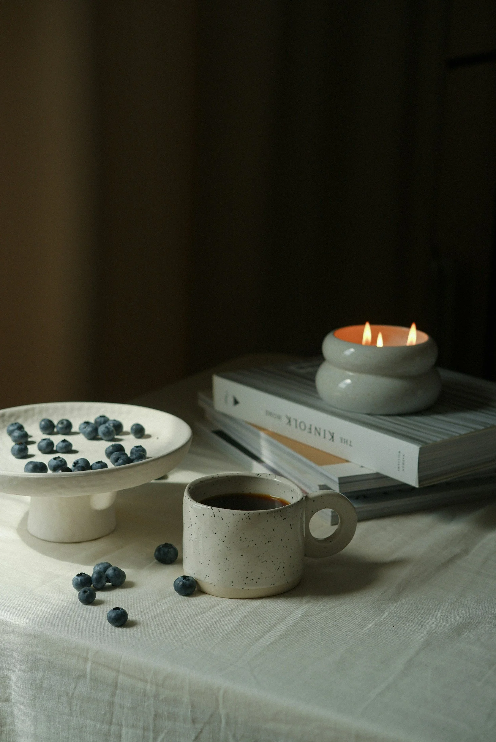 A table setup with a white tablecloth, a white mug with black speckles filled with coffee, a white pedestal dish with fresh blueberries, and a stack of books with a small candle on top, lit with a warm glow.