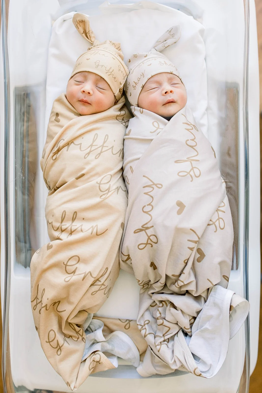 Two newborn babies swaddled in beige and white blankets with matching hats, lying side by side in a crib with clear sides and white bedding.
