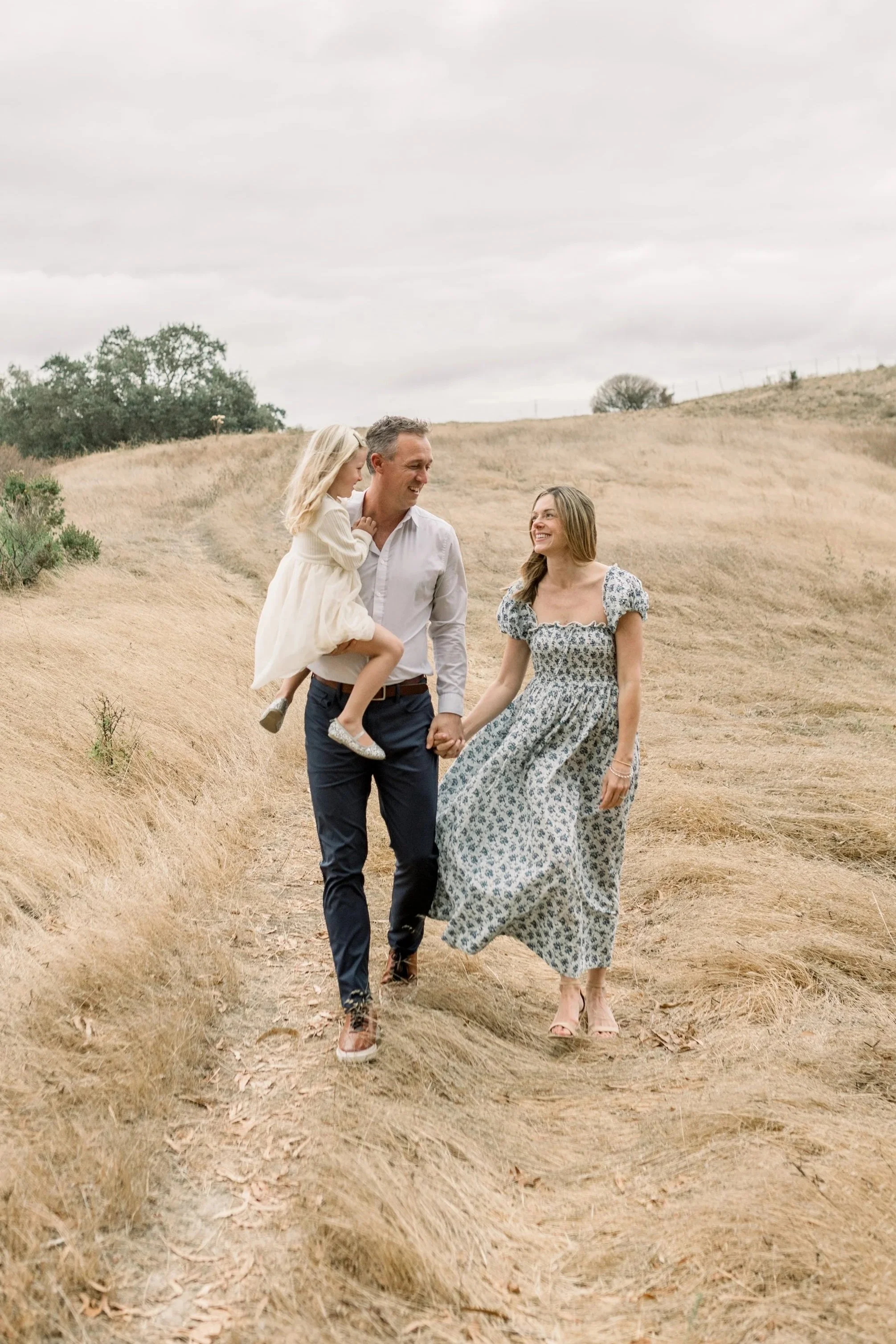 A family of three walking outdoors on a dirt path in a grassy field under a cloudy sky, with the father carrying a young girl.
