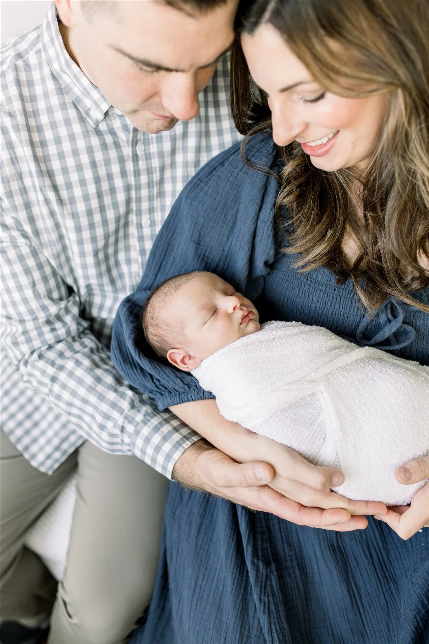Dad in checkered shirt and mom in navy dress hold their swaddled newborn son
