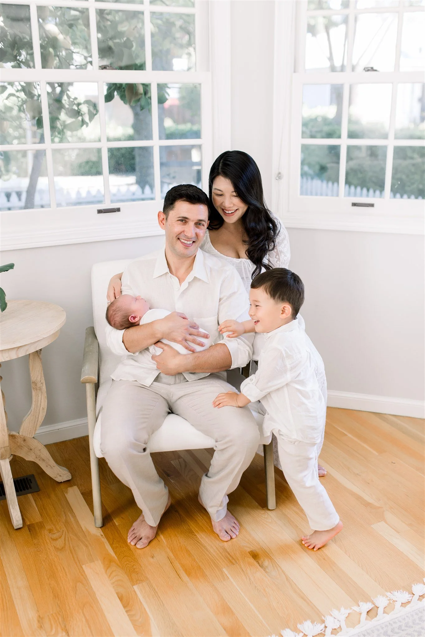 Dad sits in chair holding newborn son while other son climbs on his leg. Wife stands behind him.