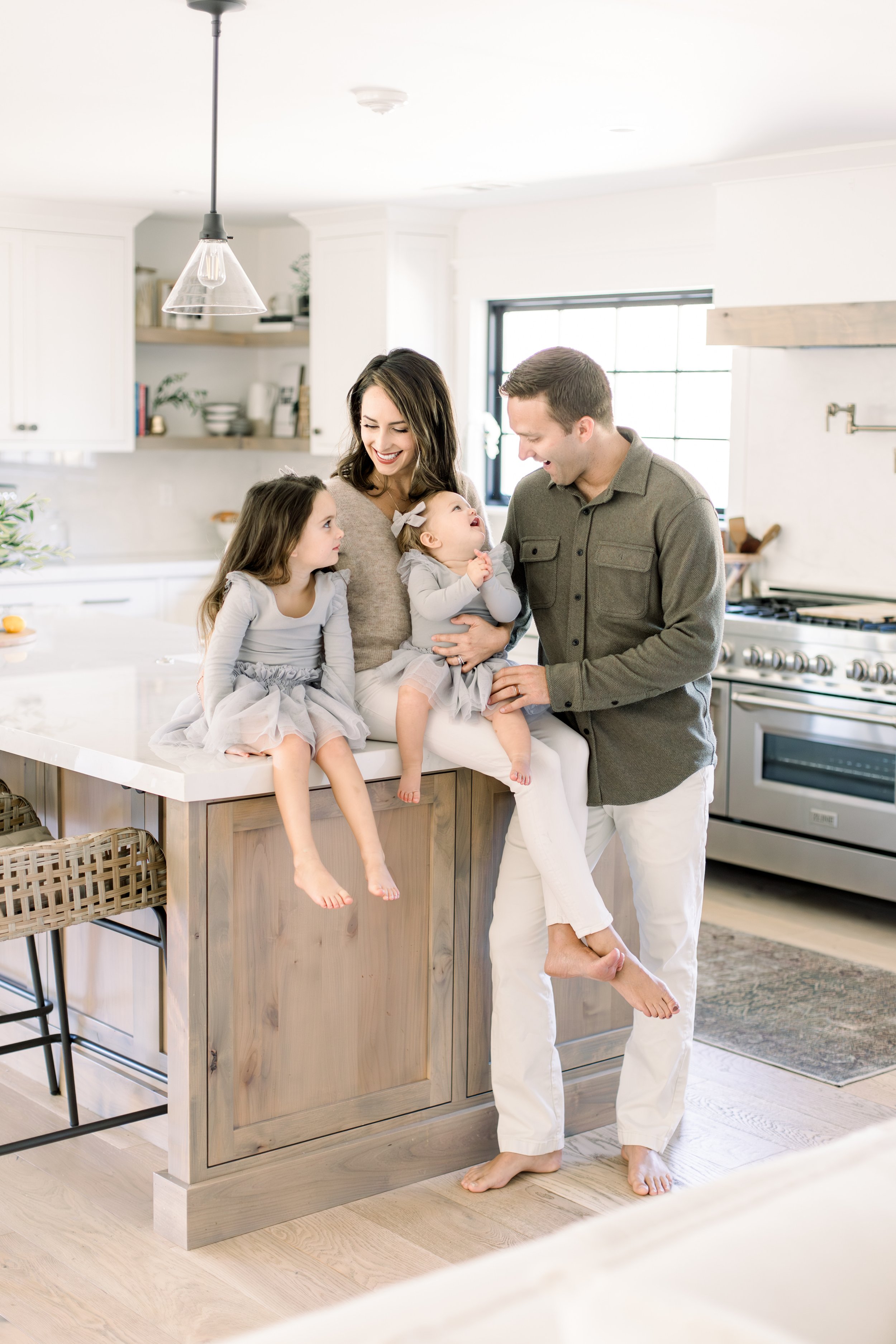A happy family of five, including two young girls, a woman, and a man, enjoying time together in a bright kitchen with white cabinets and a large window.