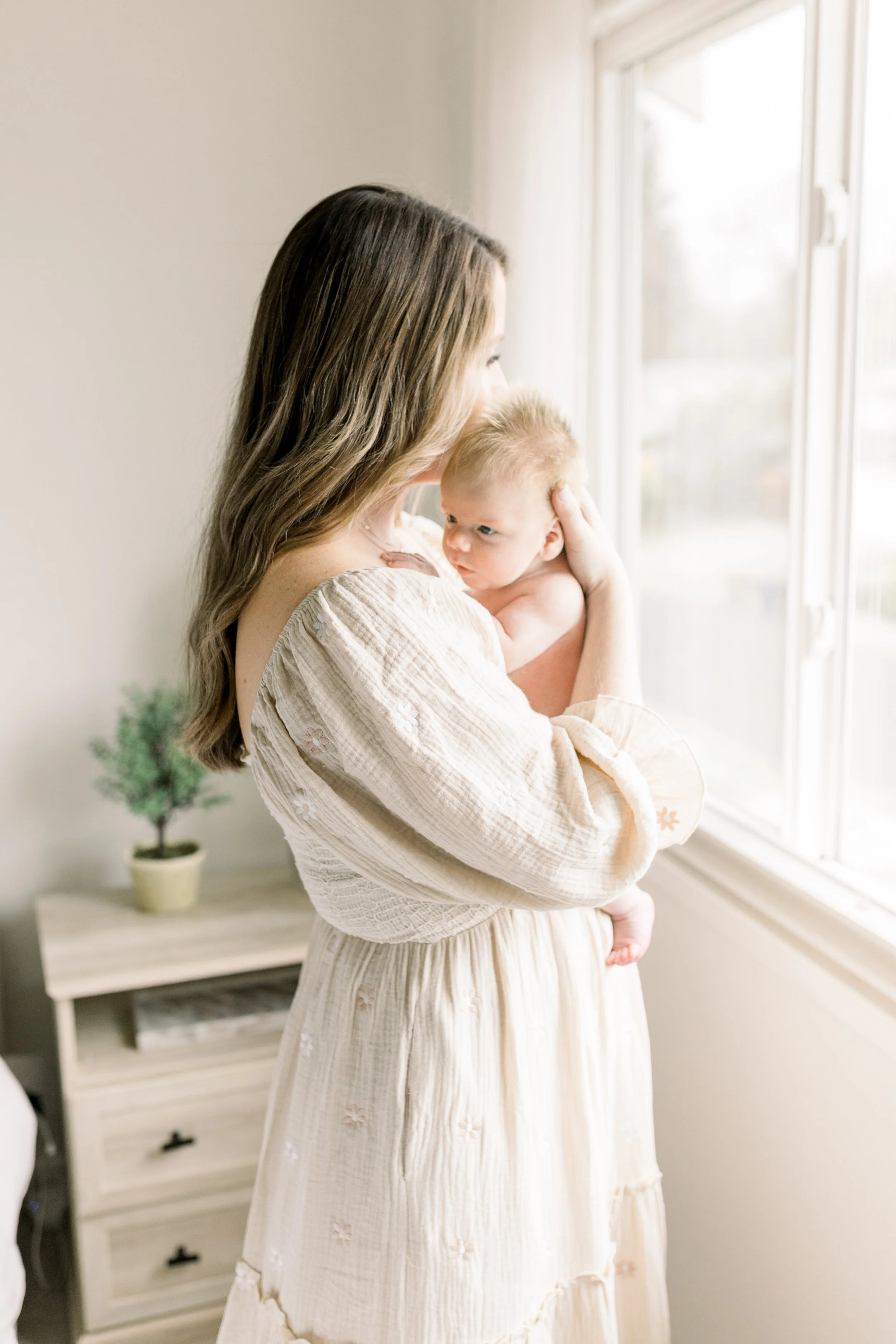 Mom in soft cream dress holding newborn son in front of window