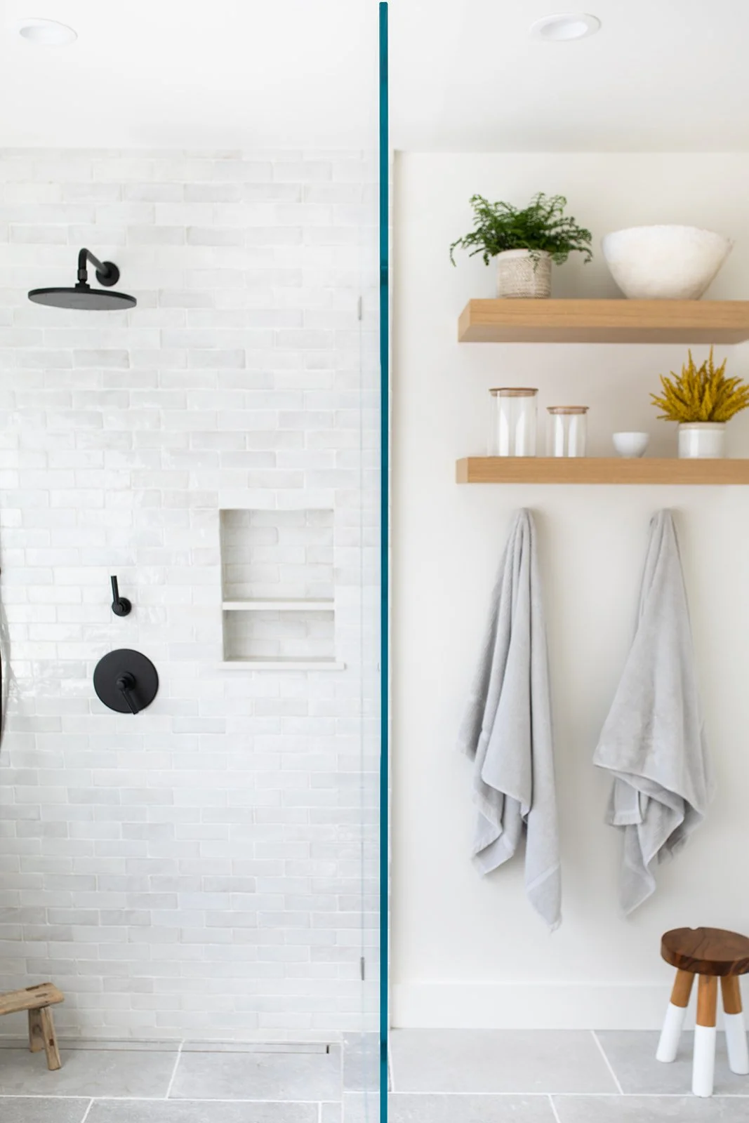 Shower stall with white tile next to wooden shelves in light and airy bathroom