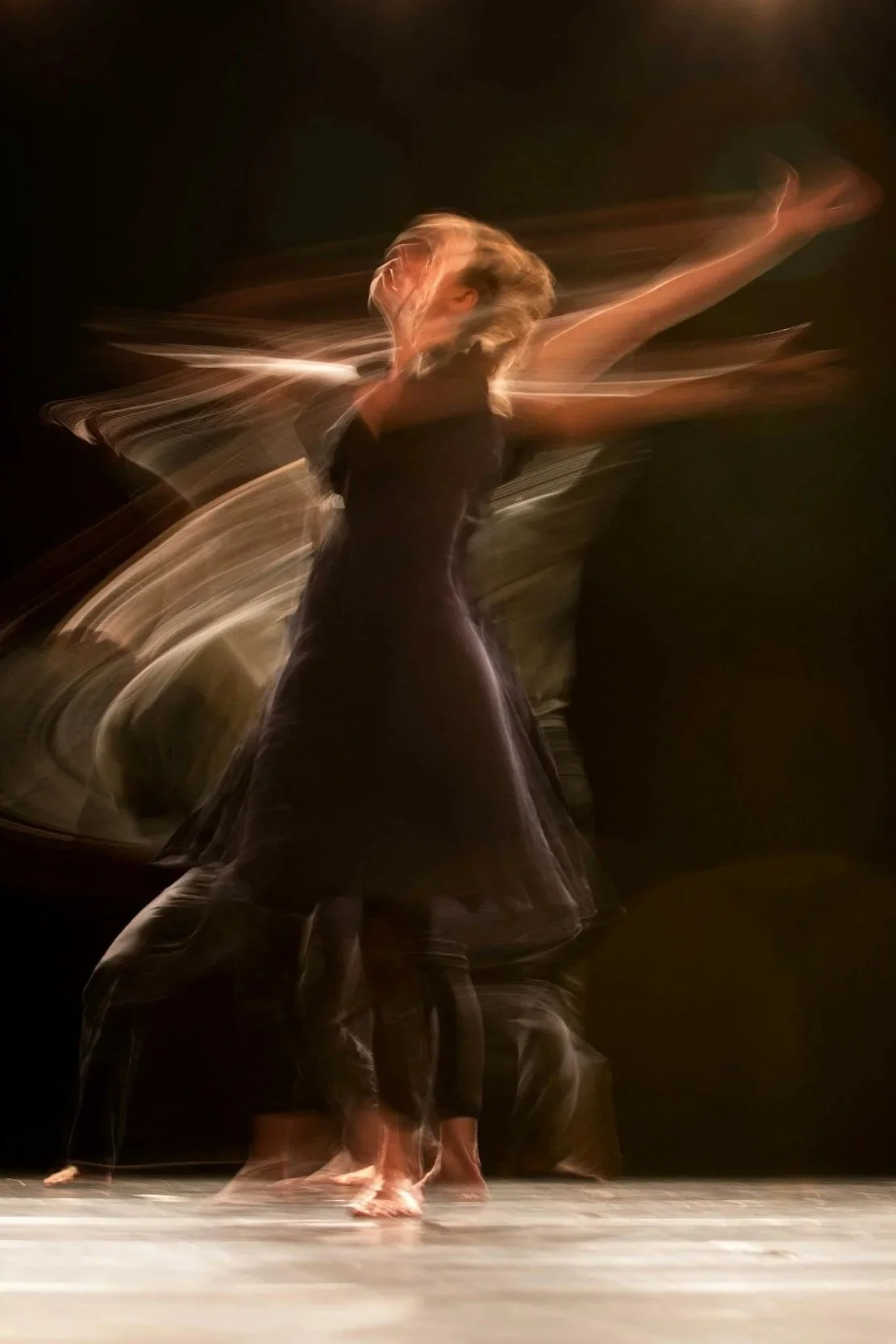 A female dancer mid-performance, captured with motion blur, wearing a black dress and standing on a stage with a dark background.