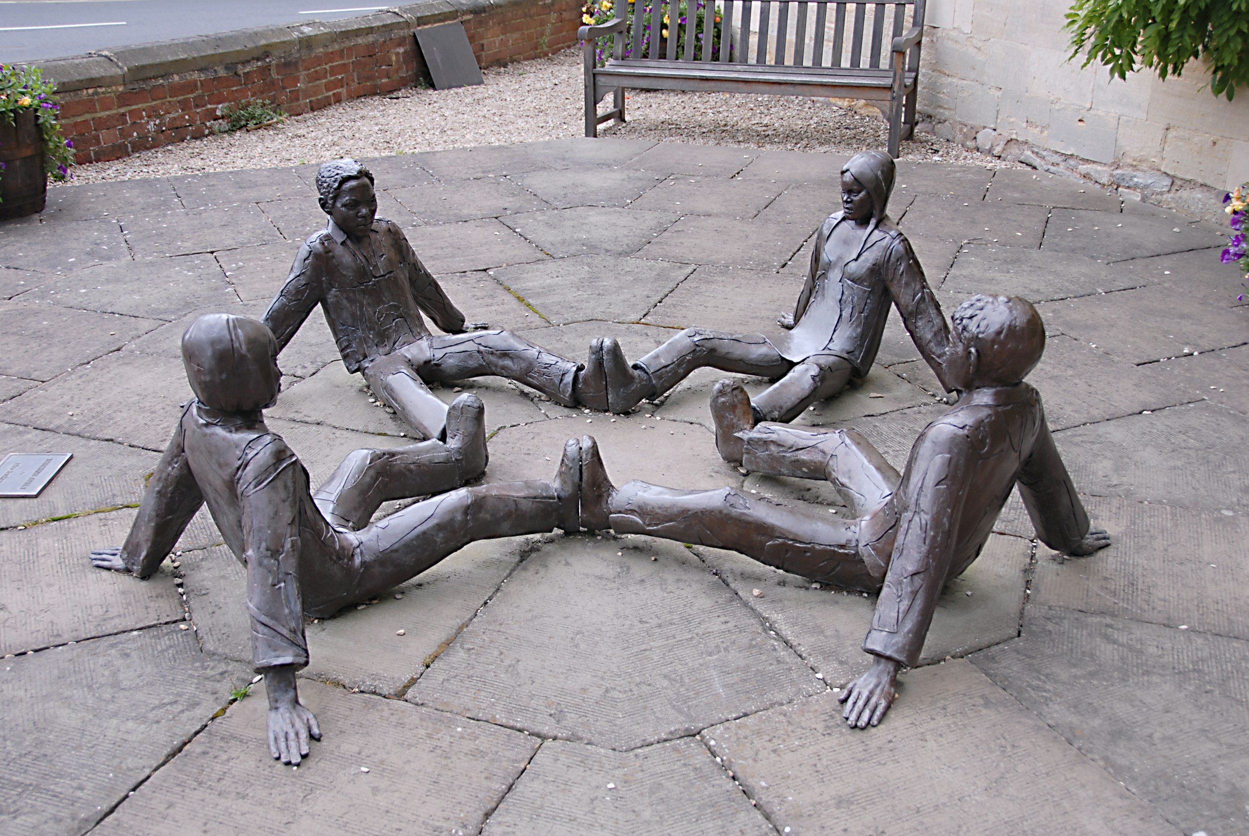 Sculpture of five people sitting in a circle with their legs stretched out, feet touching each other, located on a paved area with flowers and a bench in the background.