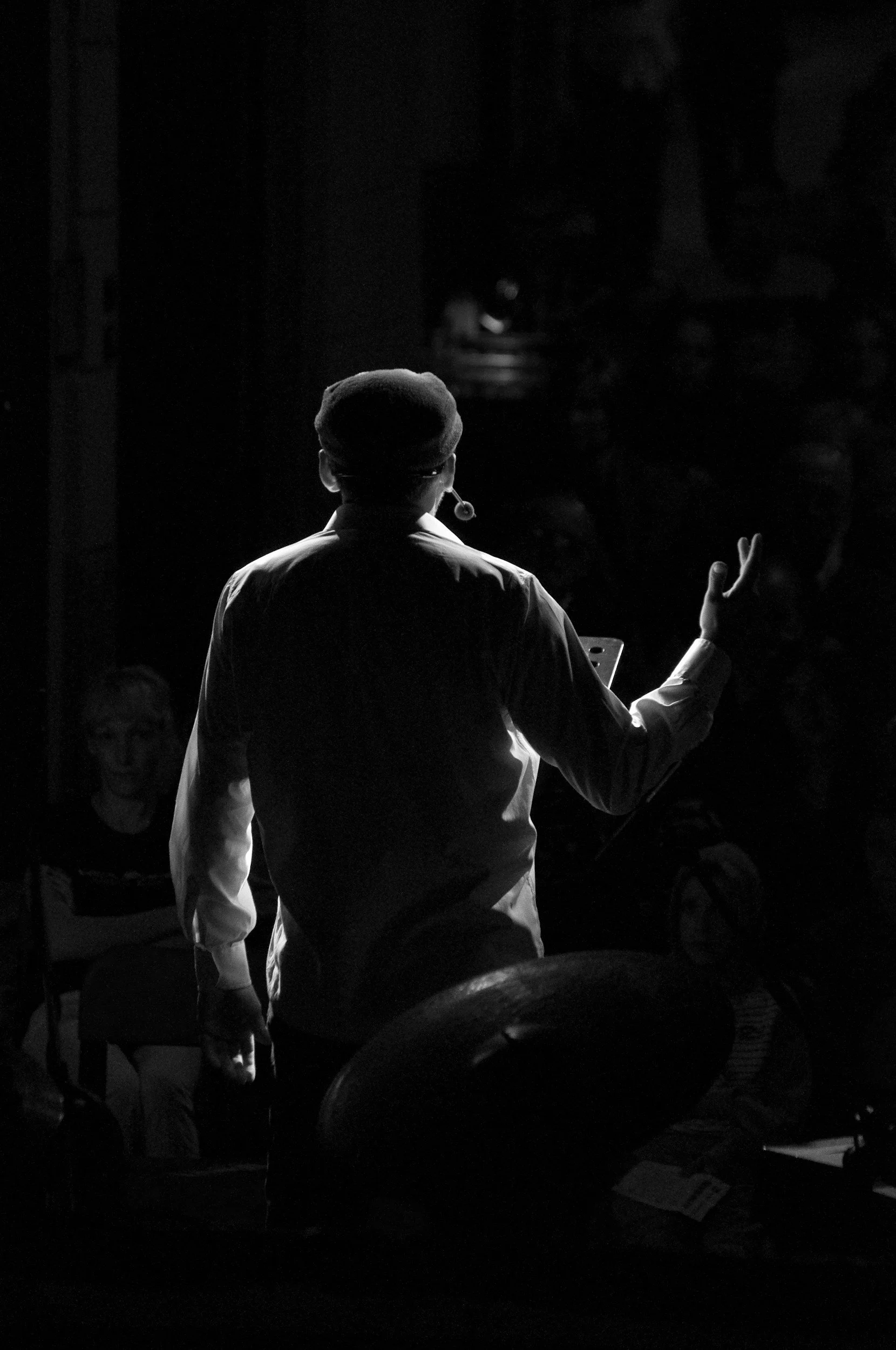 A person, seen from behind, is speaking or gesturing during a performance or event in a darkened setting, with audience members visible in the background.