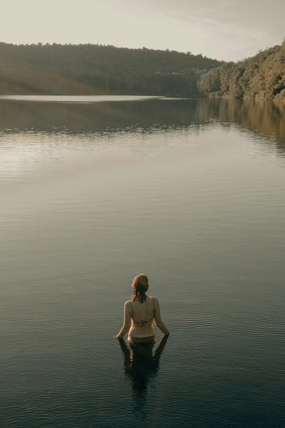 Person standing in a body of calm water during sunset, with hills in the background.