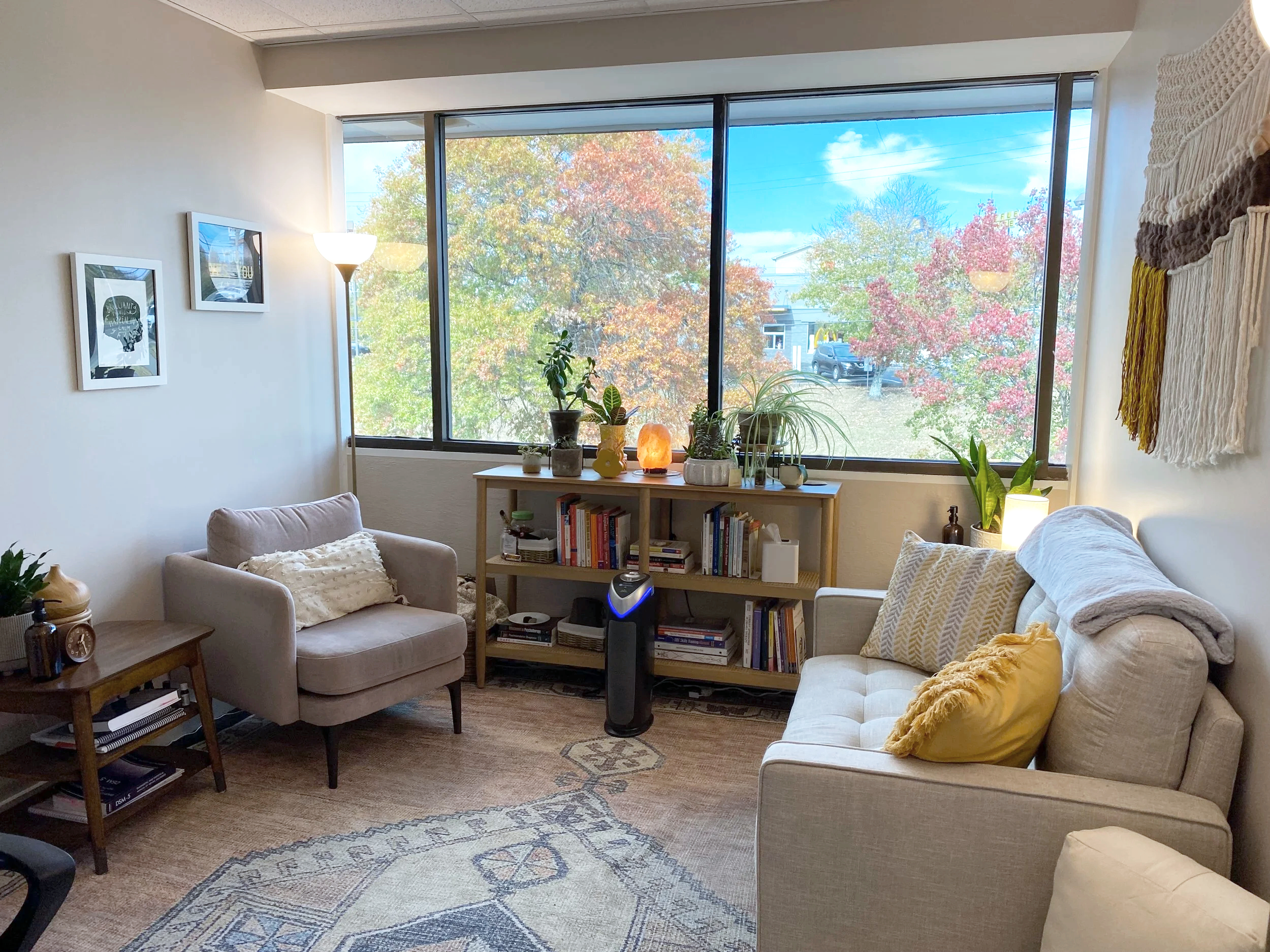 A room with a large window showing trees with colorful fall leaves. The room contains beige chairs, a wooden shelf with books and plants, and artwork on the white walls. There are various decorative items including a salt lamp, a yellow pillow, and a woven wall hanging.