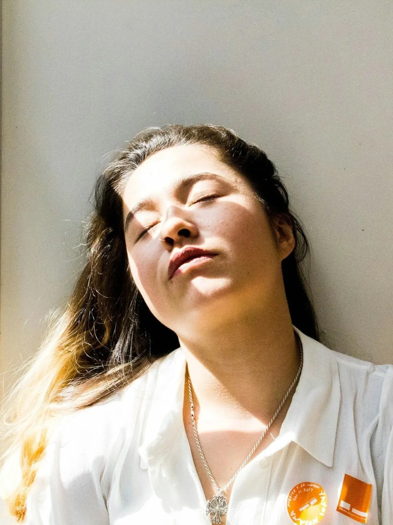 A woman with long hair, light skin, and closed eyes, looking tired in sunlight against a plain wall, wearing a white shirt and a necklace.