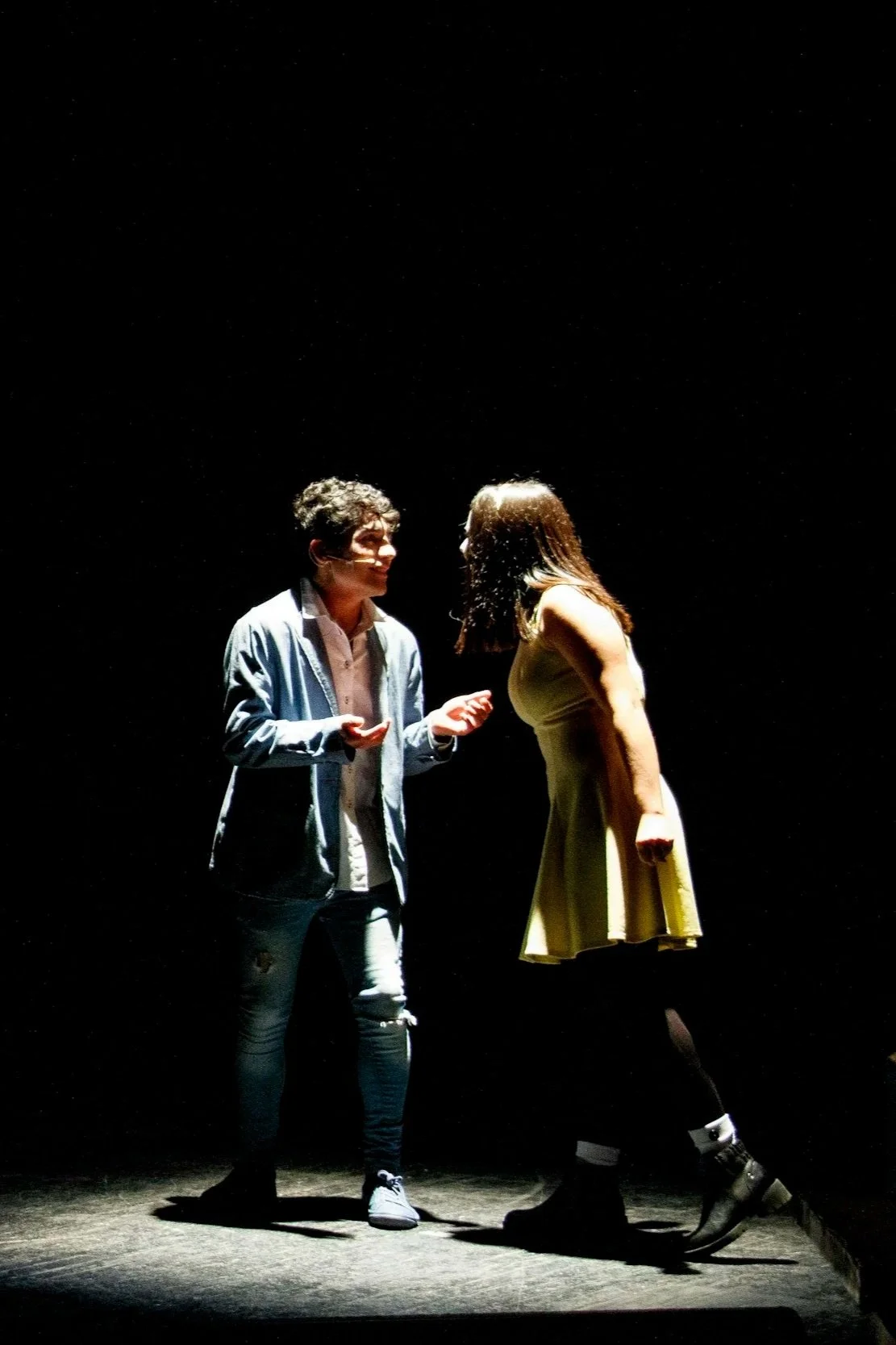 Two people, a man and a woman, are standing on a dark stage with spotlight illumination. They are facing each other, engaged in conversation or performance.