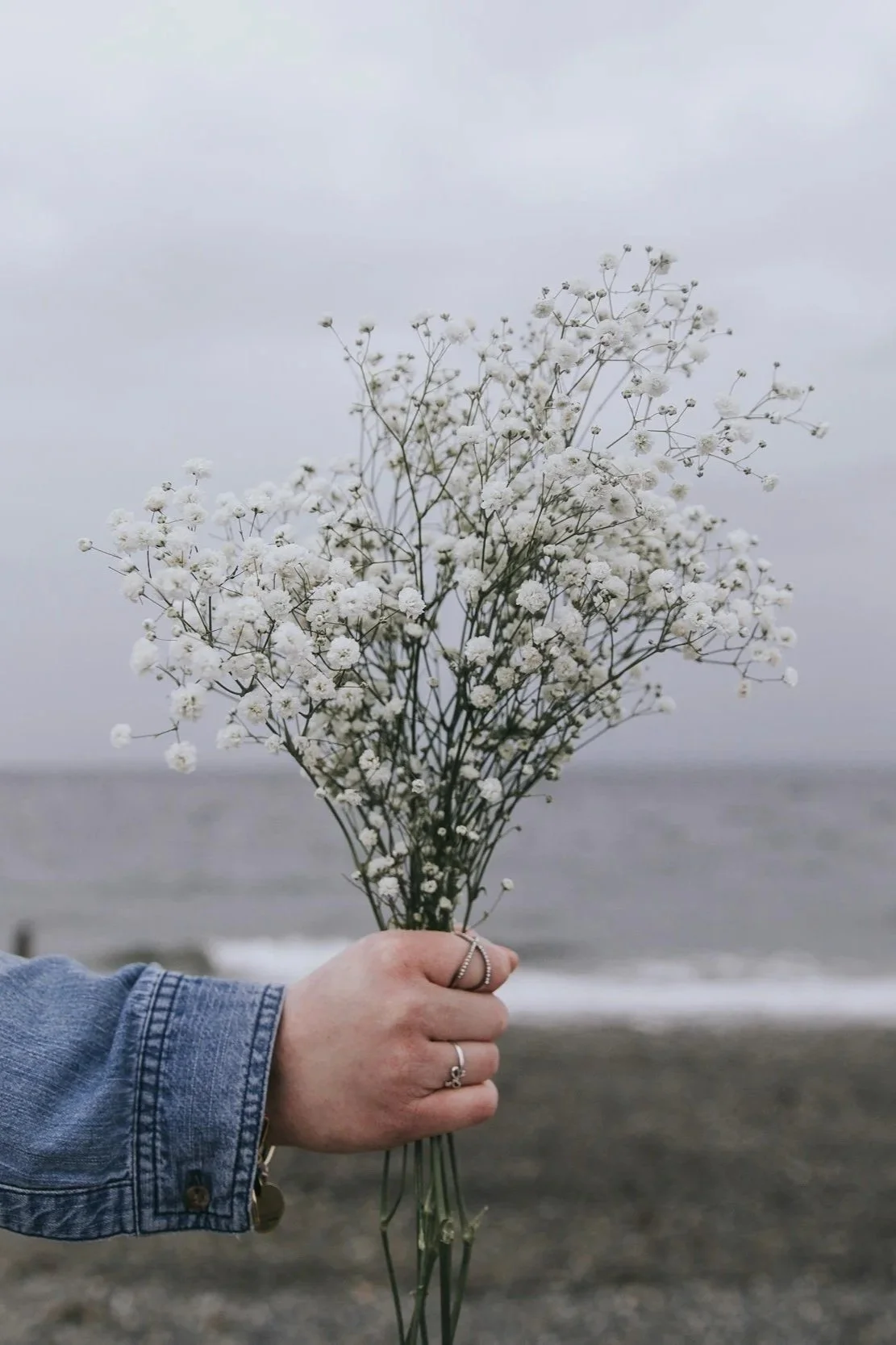 A person holding a bouquet of white baby's breath flowers against a cloudy sky and a distant body of water.