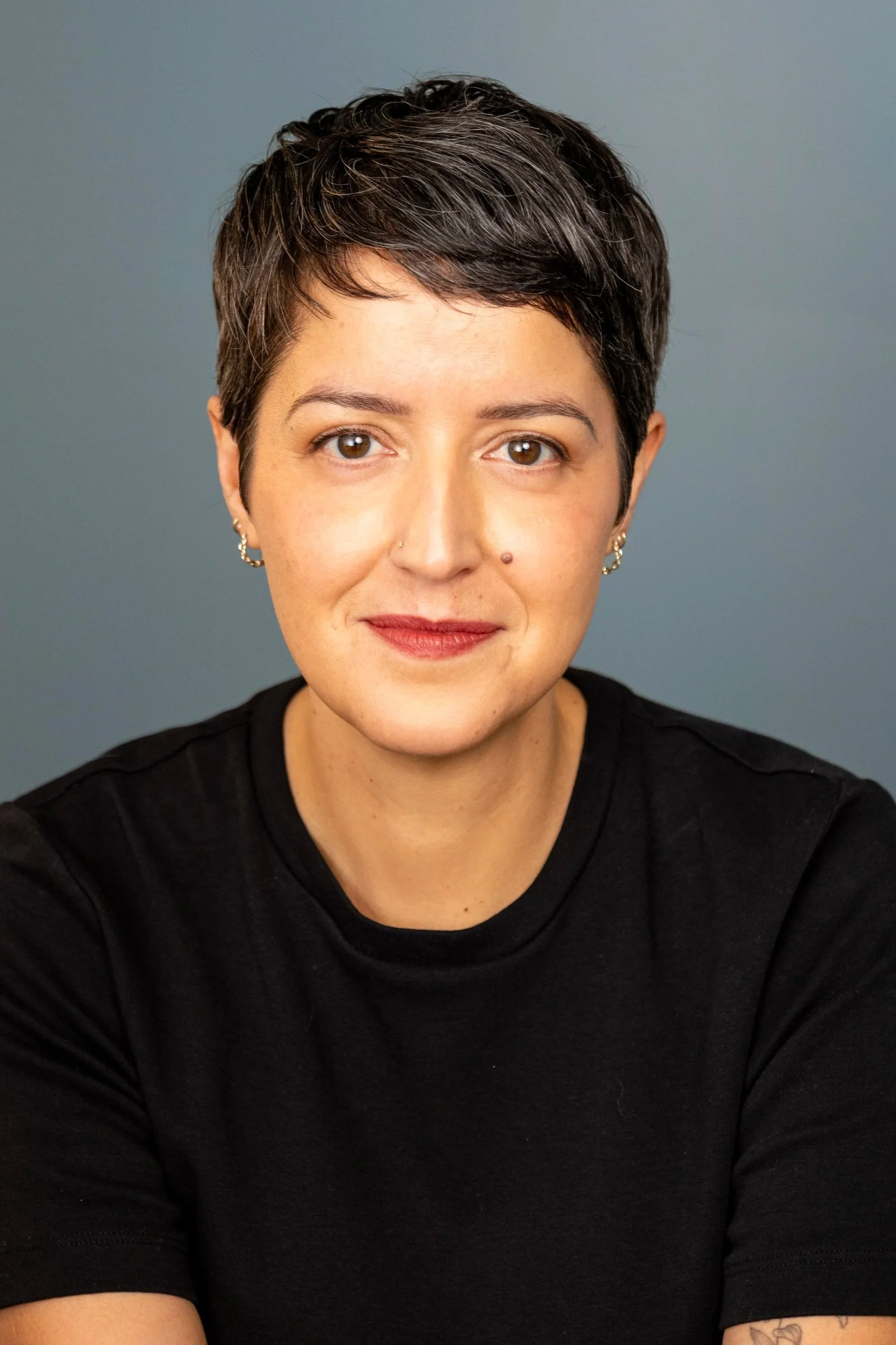 Close-up front view of a woman with short dark hair, brown eyes, light skin, wearing small earrings and a black top, smiling slightly, against a plain gray background.