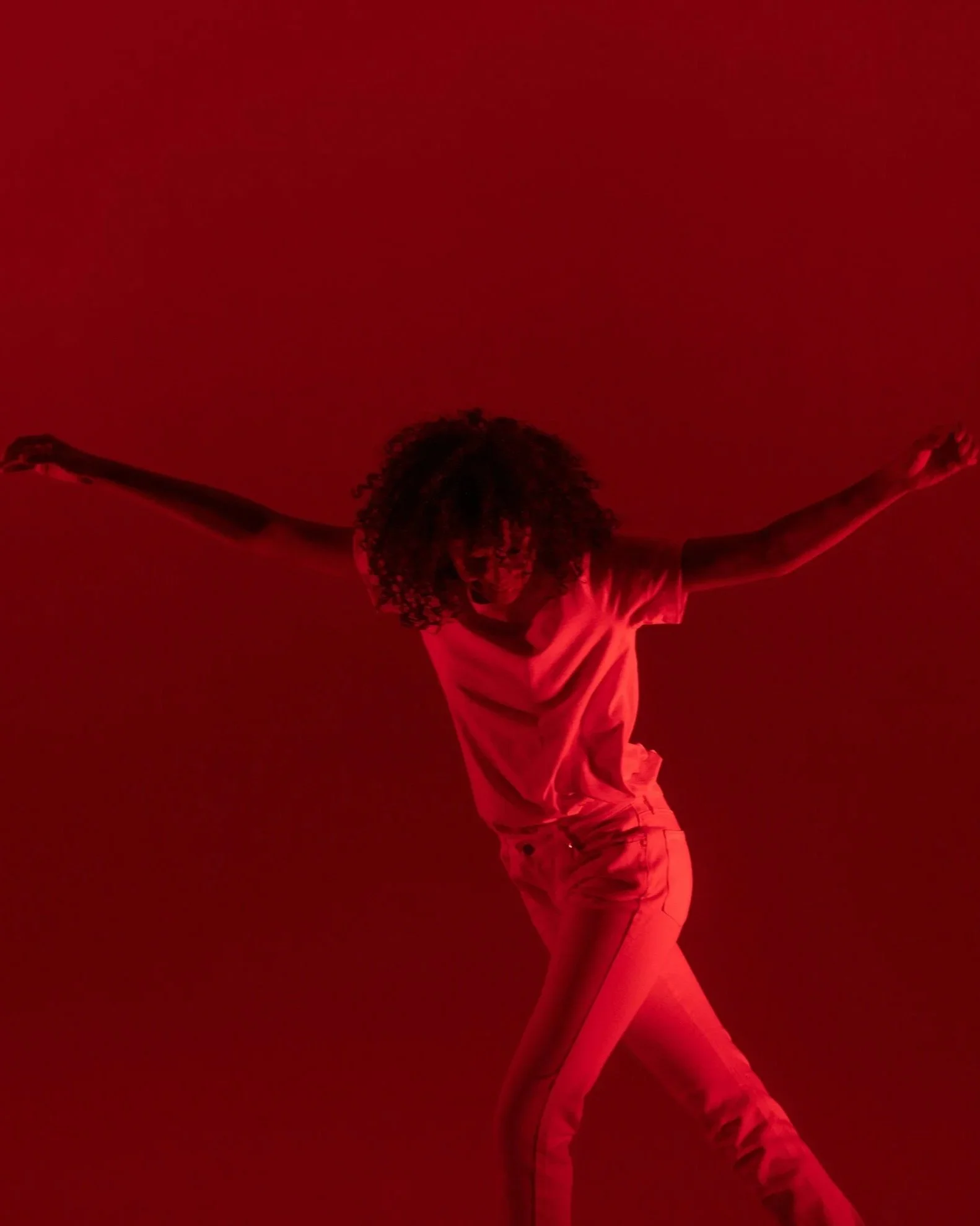 A person with curly hair wearing casual clothing is in a physical posture, looking down, against a red background with arms extended.