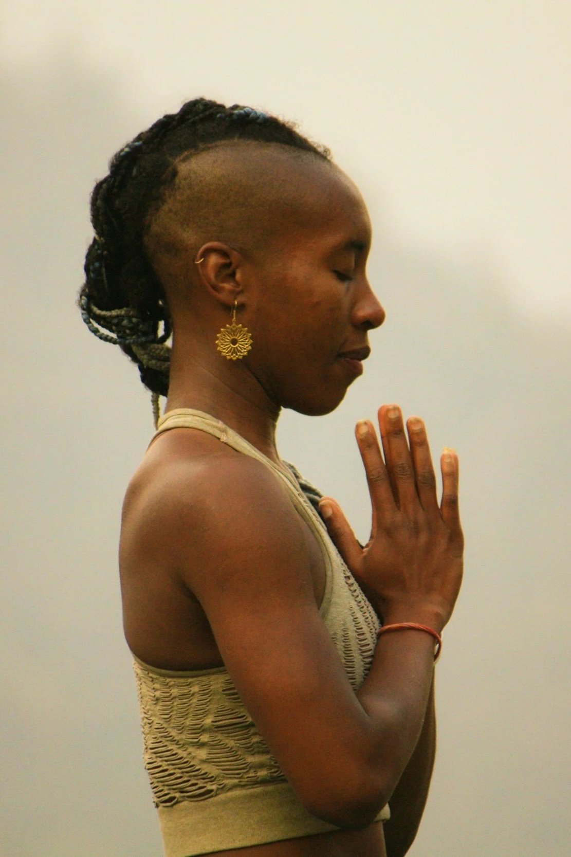 A woman with braided hair in a bun, wearing a sleeveless top, gold earring, and a bracelet, practicing yoga or meditation with eyes closed and hands in a prayer position.