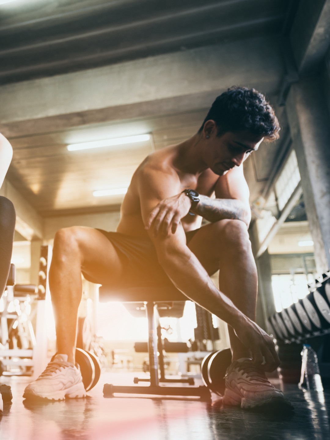 A young man in a gym adjusting his shoe while sitting on a workout bench with dumbbells around him.