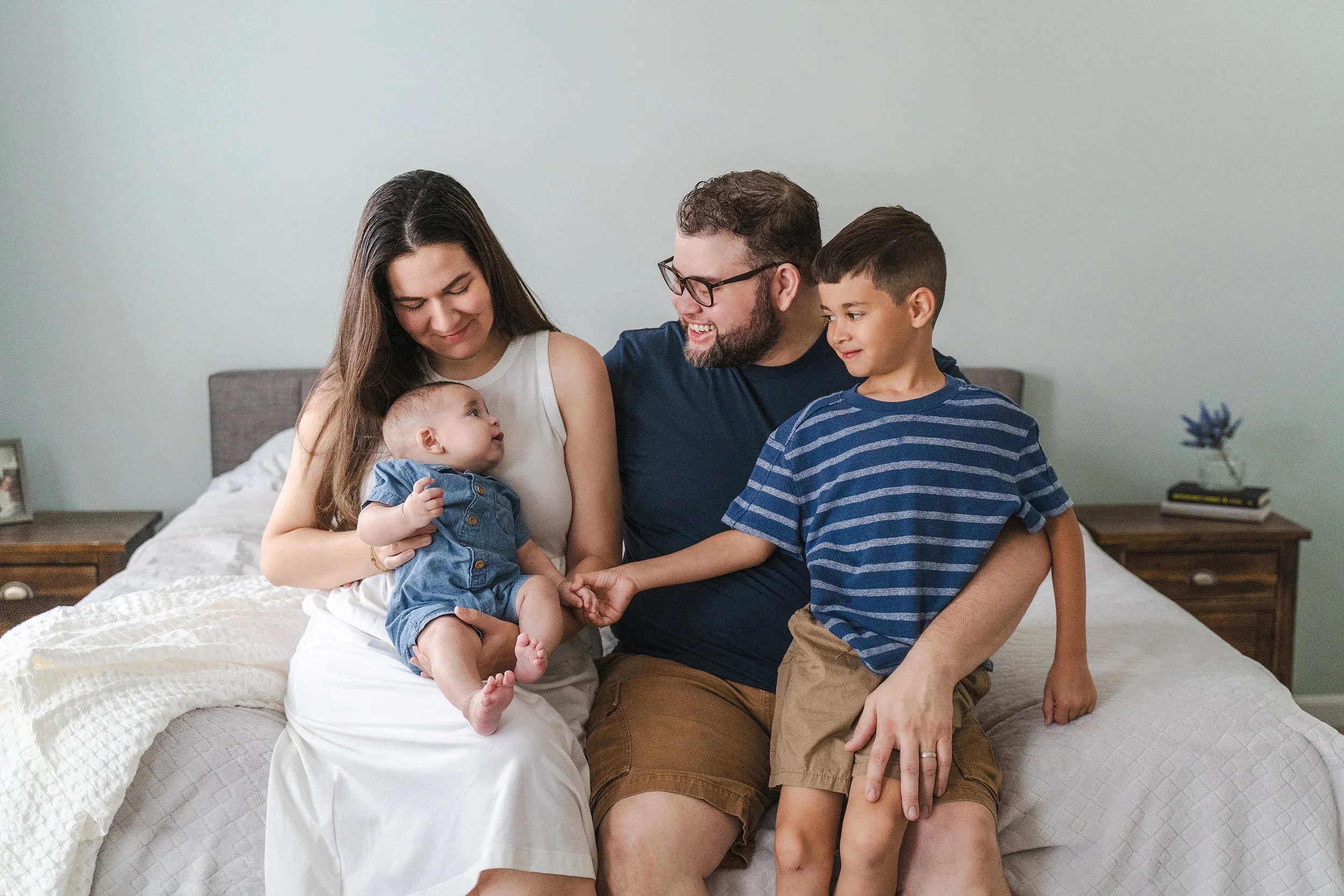 Family of four sits on mom and dad's bed together looking at and playing with their new baby in their home in Clarksville, TN.