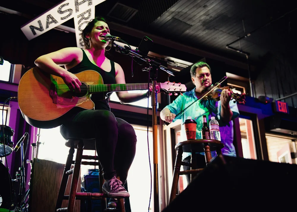 A singer with a guitar and a fiddler performing on a small stage