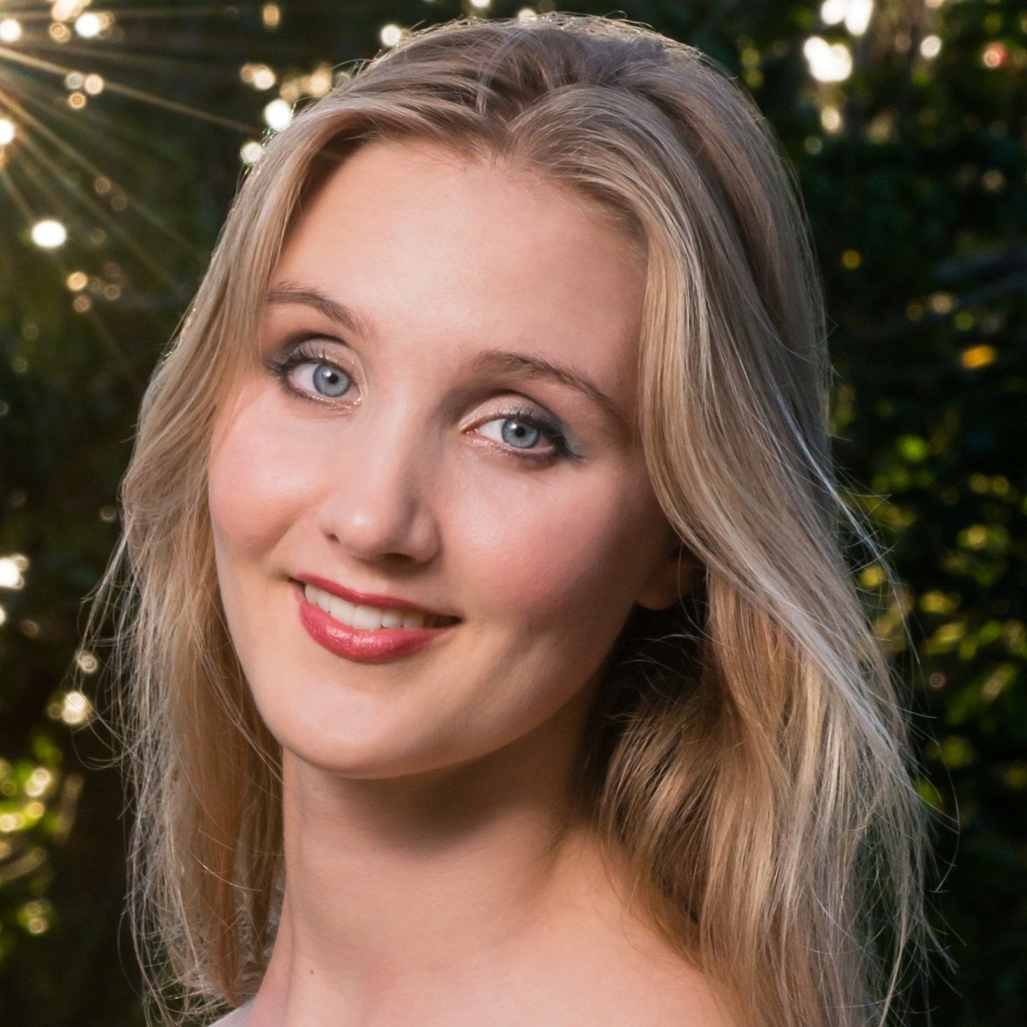 A young woman with blonde hair and blue eyes smiling outdoors with sunlight filtering through trees in the background.