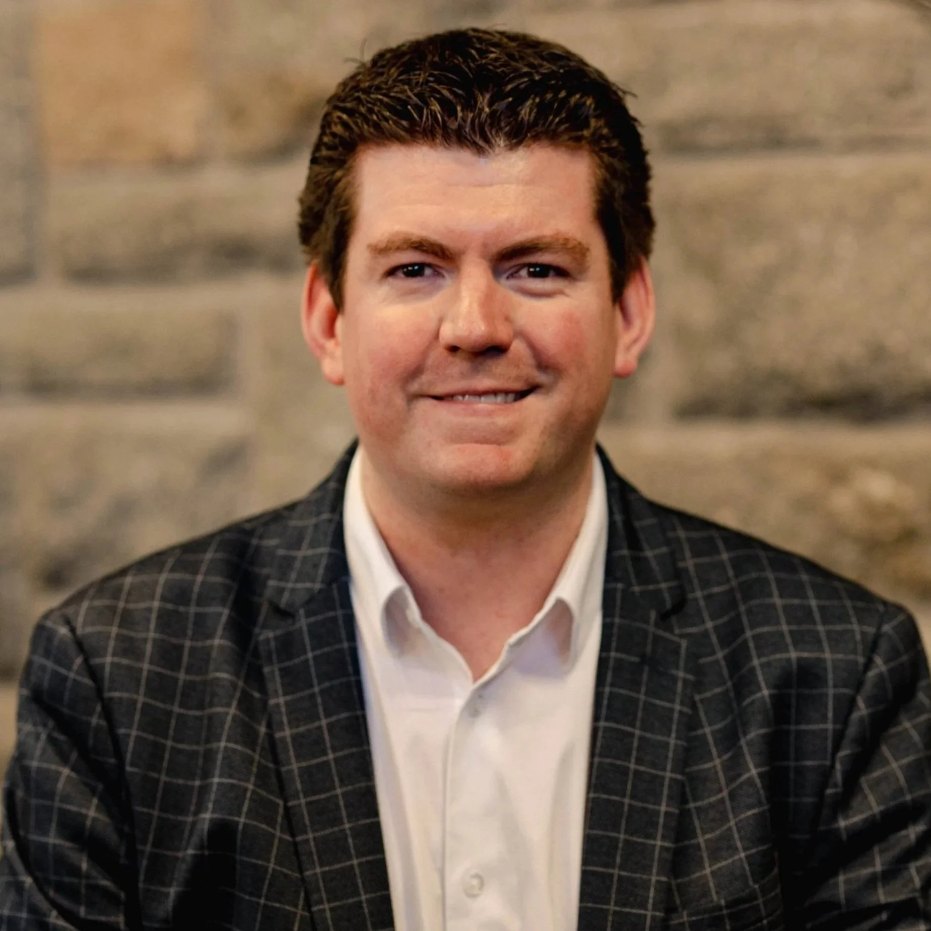 A man with short brown hair, wearing a black checkered blazer and white shirt, smiling in front of a stone wall background.