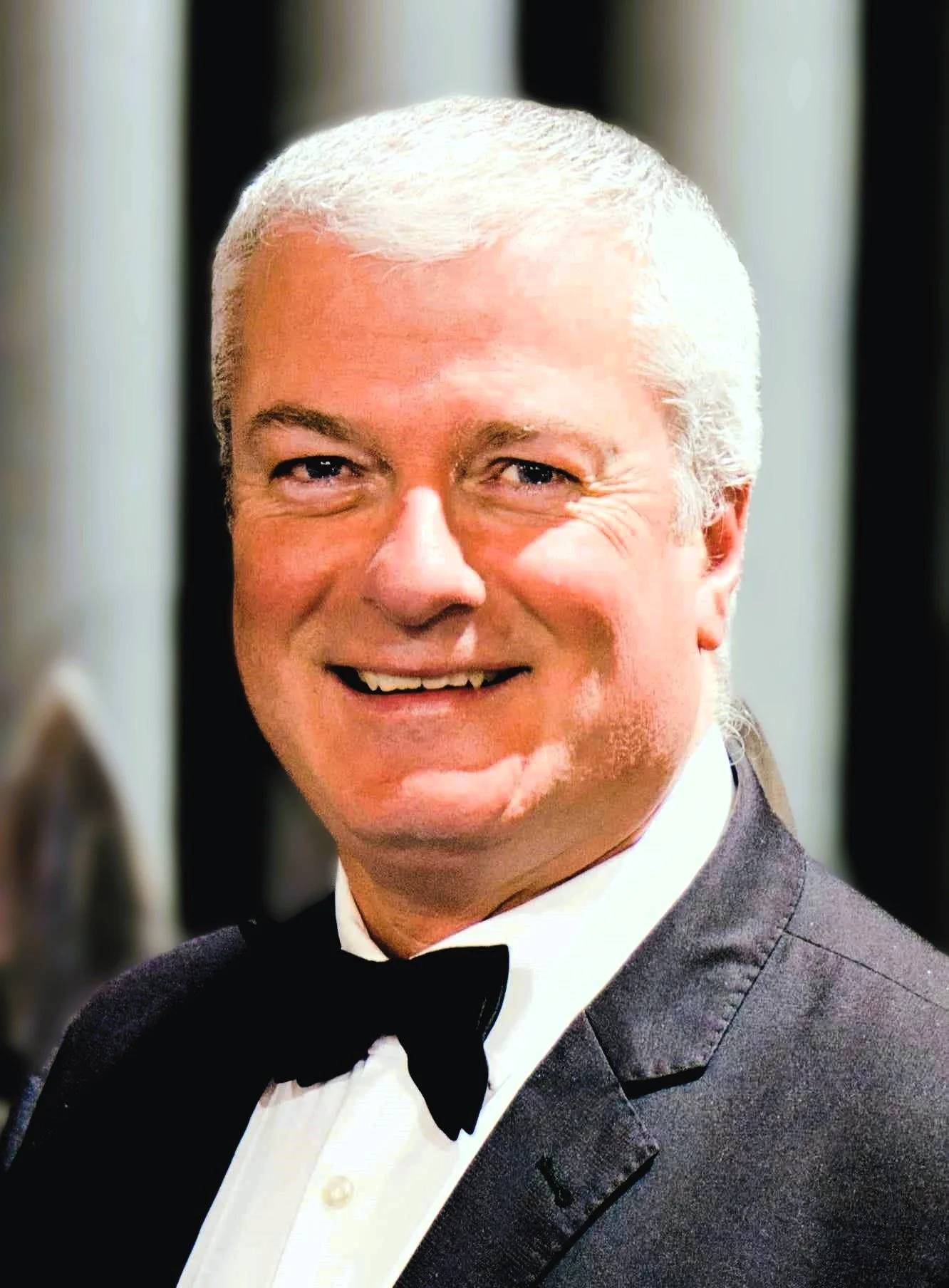 A smiling man with gray hair, wearing a tuxedo with a bow tie, at a formal event.