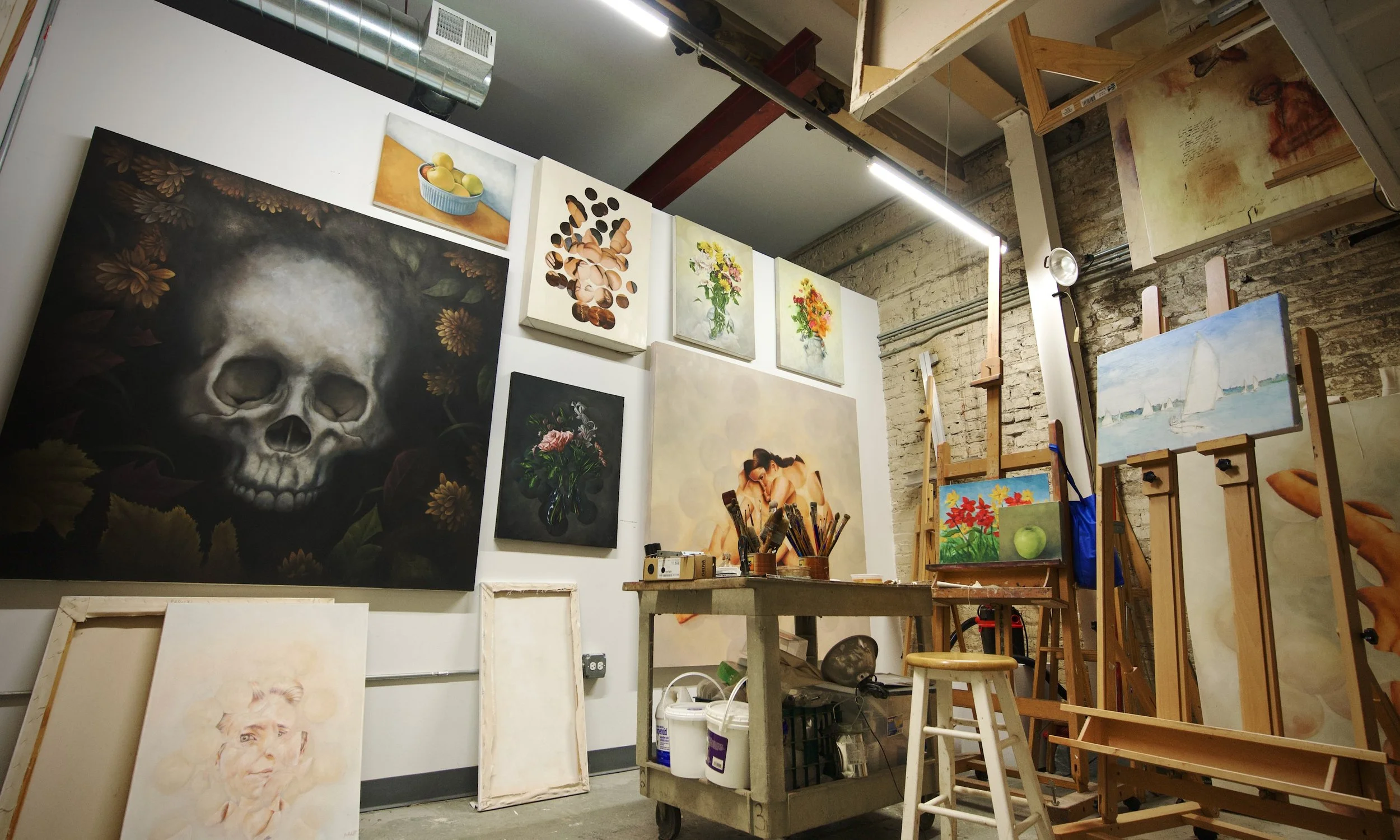 An art studio with an assortment of paintings hanging along walls, several easels – some with paintings on them – and a cart with painting supplies and brushes in the foreground