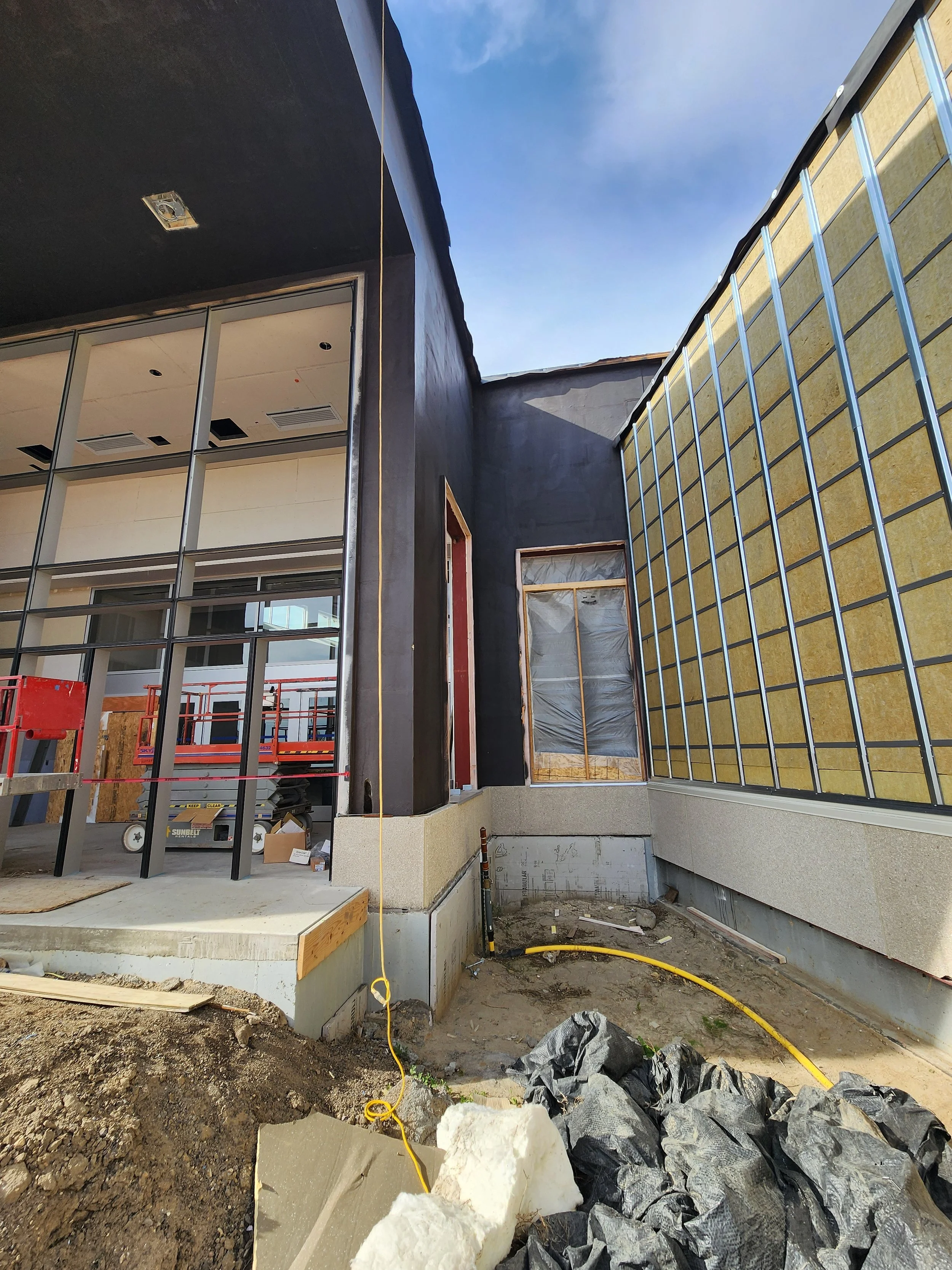 Construction site with exterior of a building under renovation, showing walls with insulation and framing, construction materials, and equipment on dirt ground.