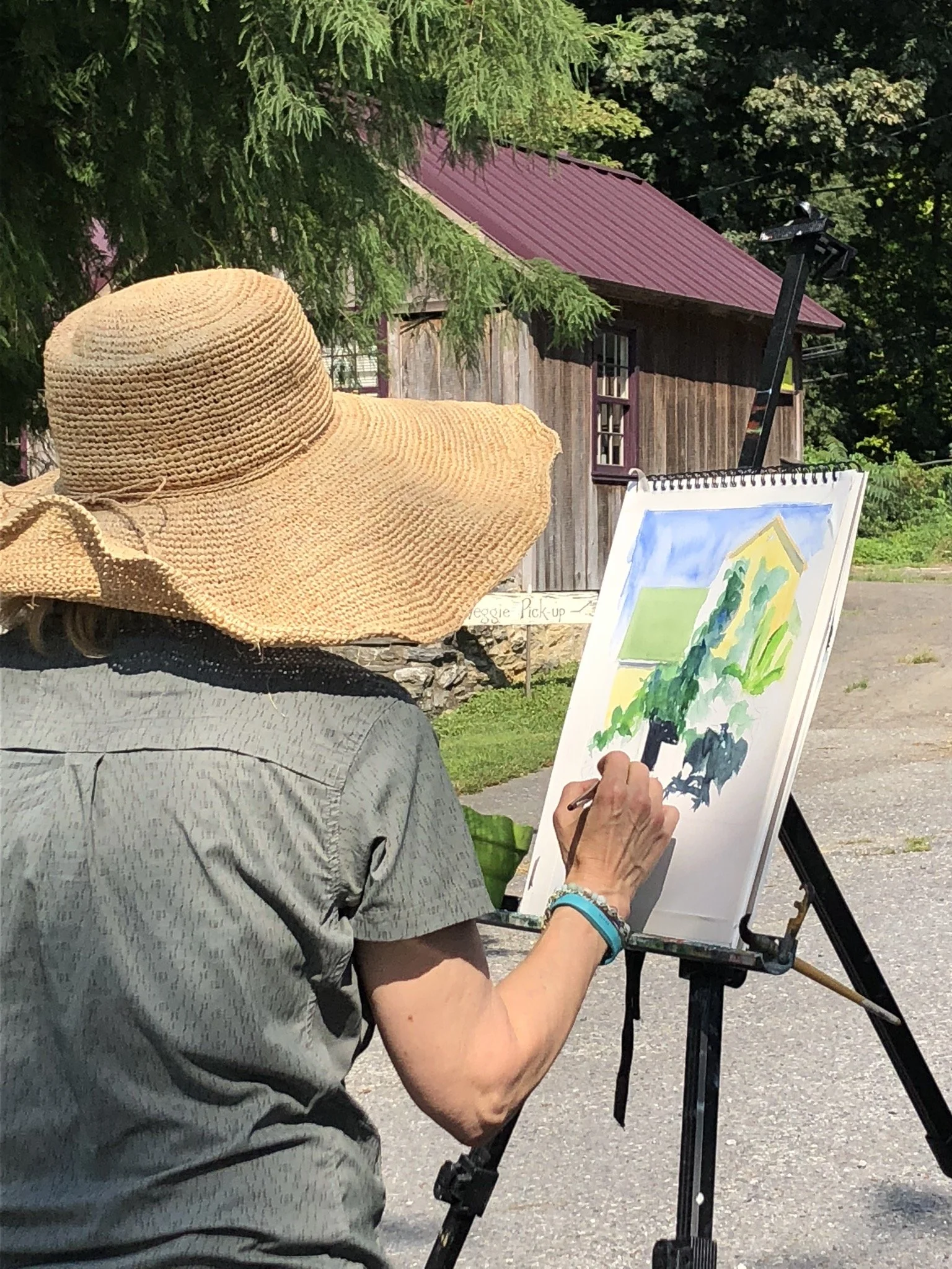 A woman in a large straw hat in front of an easel, watercoloring a landscape.