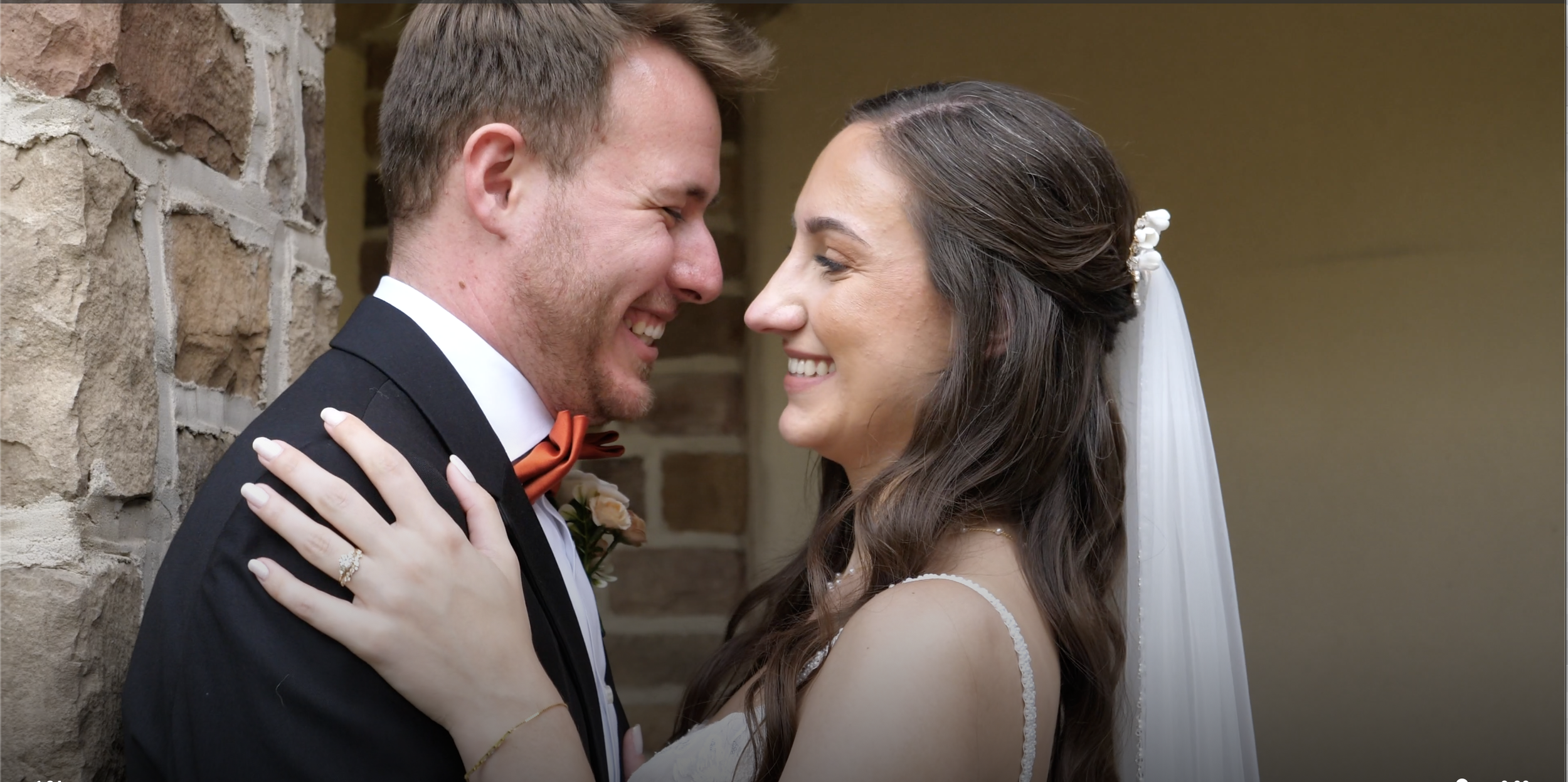 A bride and groom face each other smiling outdoors on a wedding day, with green trees in the background. The bride wears a white lace wedding dress and holds a colorful bouquet, while the groom wears a blue suit with a tie and boutonniere.