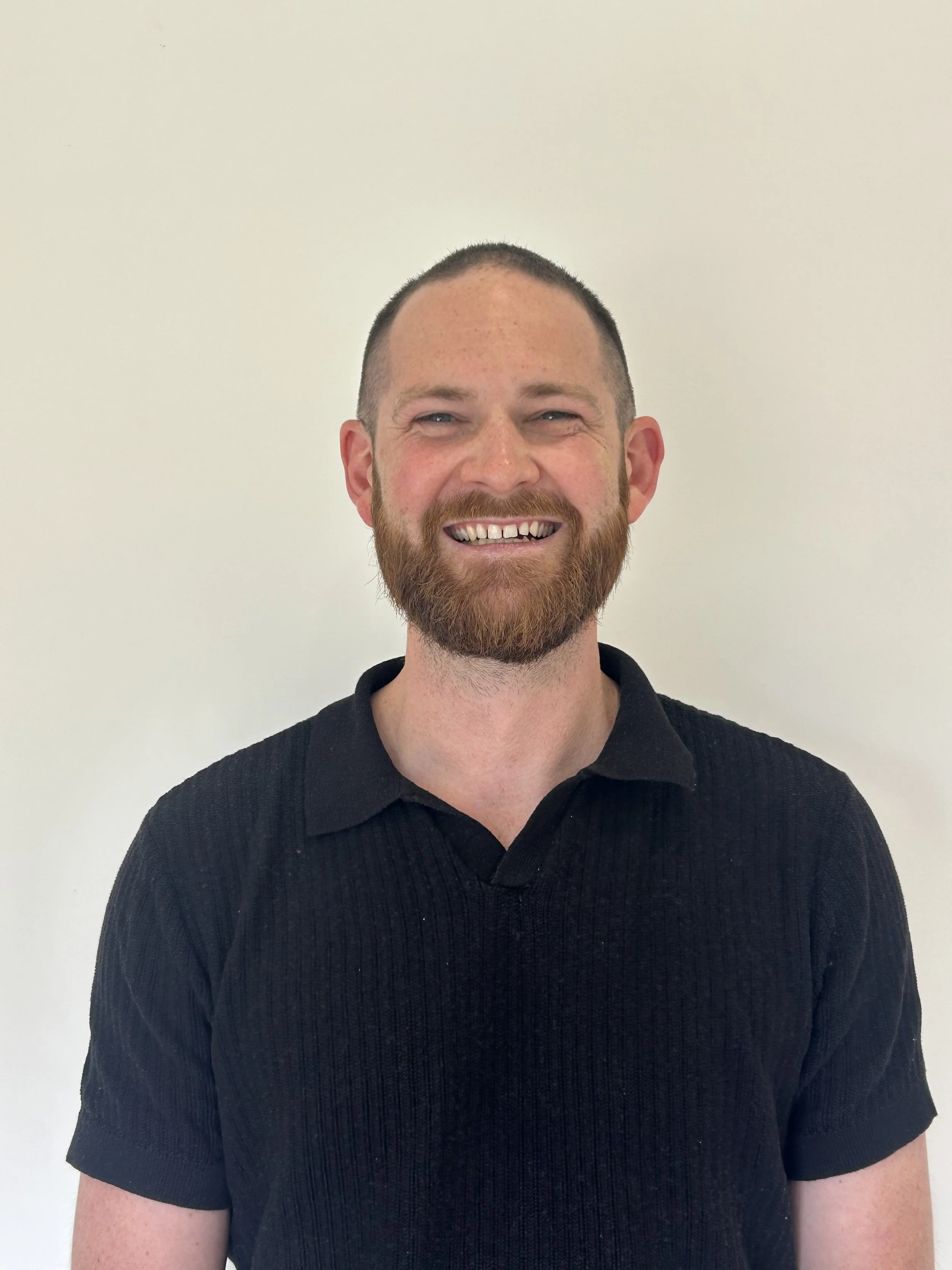 A smiling man with a beard and short hair, wearing a black polo shirt, standing against a plain white wall.