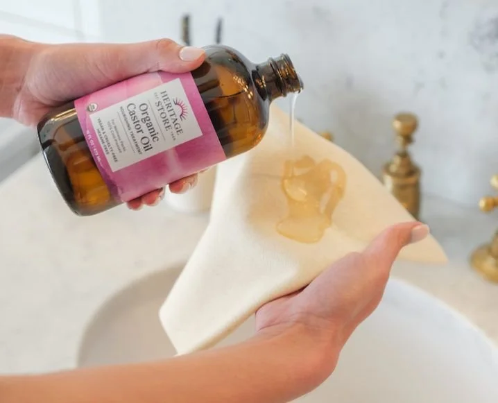 A person pours castor oil from a brown bottle onto a white cloth in a bathroom with a sink and gold fixtures in the background.